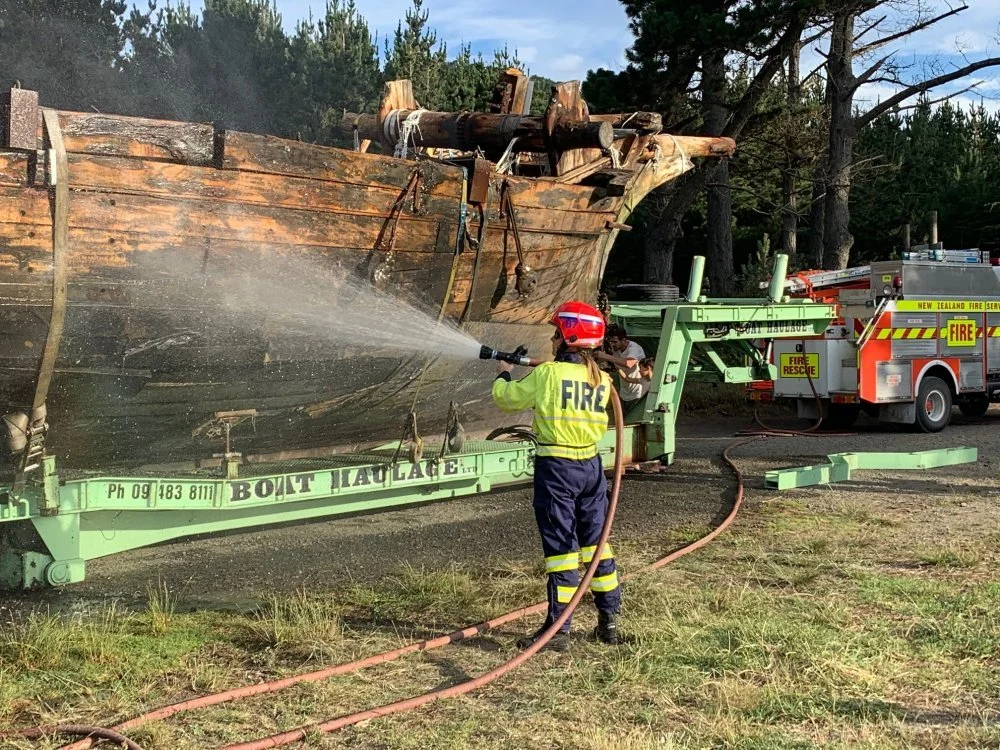 A firefighter in yellow gear sprays water on a large wooden boat that is on a green trailer, with a fire truck in the background.