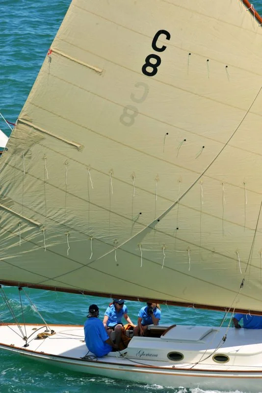 Sight of a sailboat with three crew members, navigating on the ocean under a large beige sail marked with a black number 8 and identifiable 'C' shape, with a visible blue sky and water.