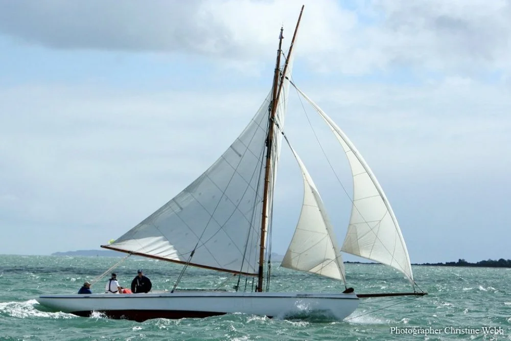 A sailboat with white sails on the water with three people on board, under a cloudy sky.