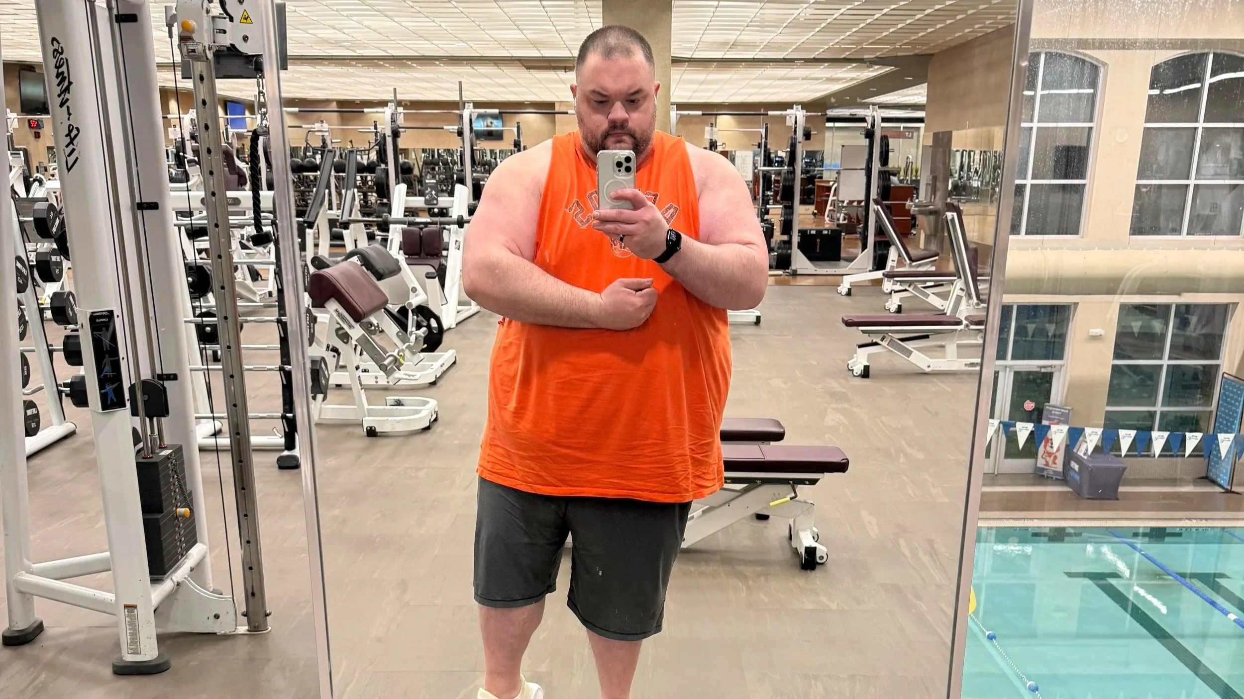 A man taking a selfie in a gym mirror. He is wearing a bright orange sleeveless shirt, black shorts, and a smartwatch. The gym has exercise equipment in the background.