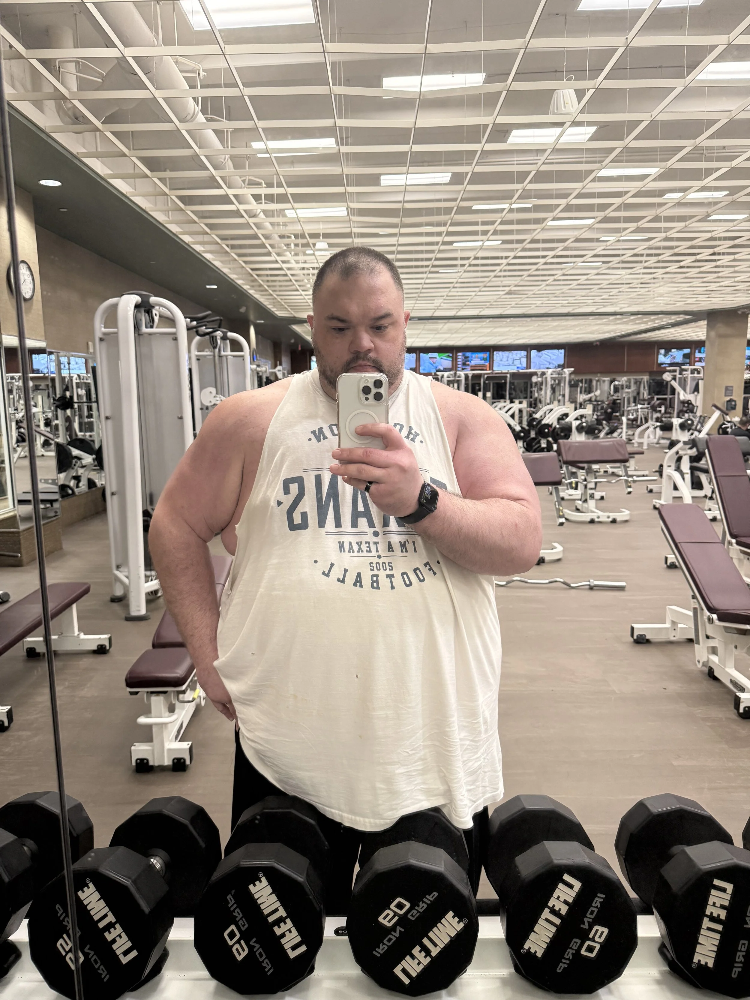 A man taking a mirror selfie in a gym, wearing a sleeveless shirt, with gym equipment and dumbbells visible in the background.