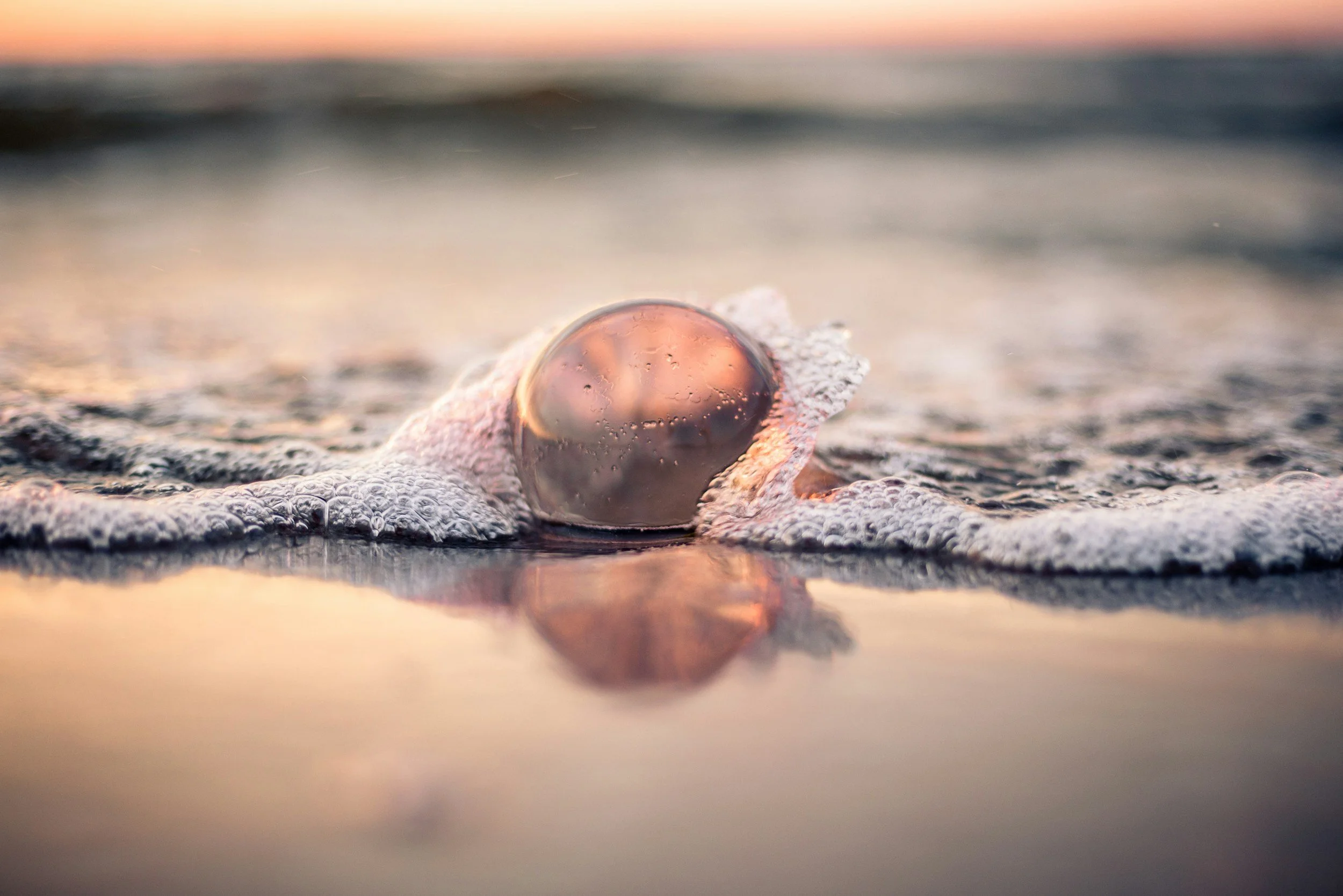 Eine goldene Perle, von einer Meerwelle sanft an den Strand gespült, während das Wasser im Hintergrund bei Sonnenaufgang reflektiert. Eine Szenerie, die die pure Essenz unseres Seins repräsentiert, in Ruhe, Schönheit, Frieden und Geborgenheit.
