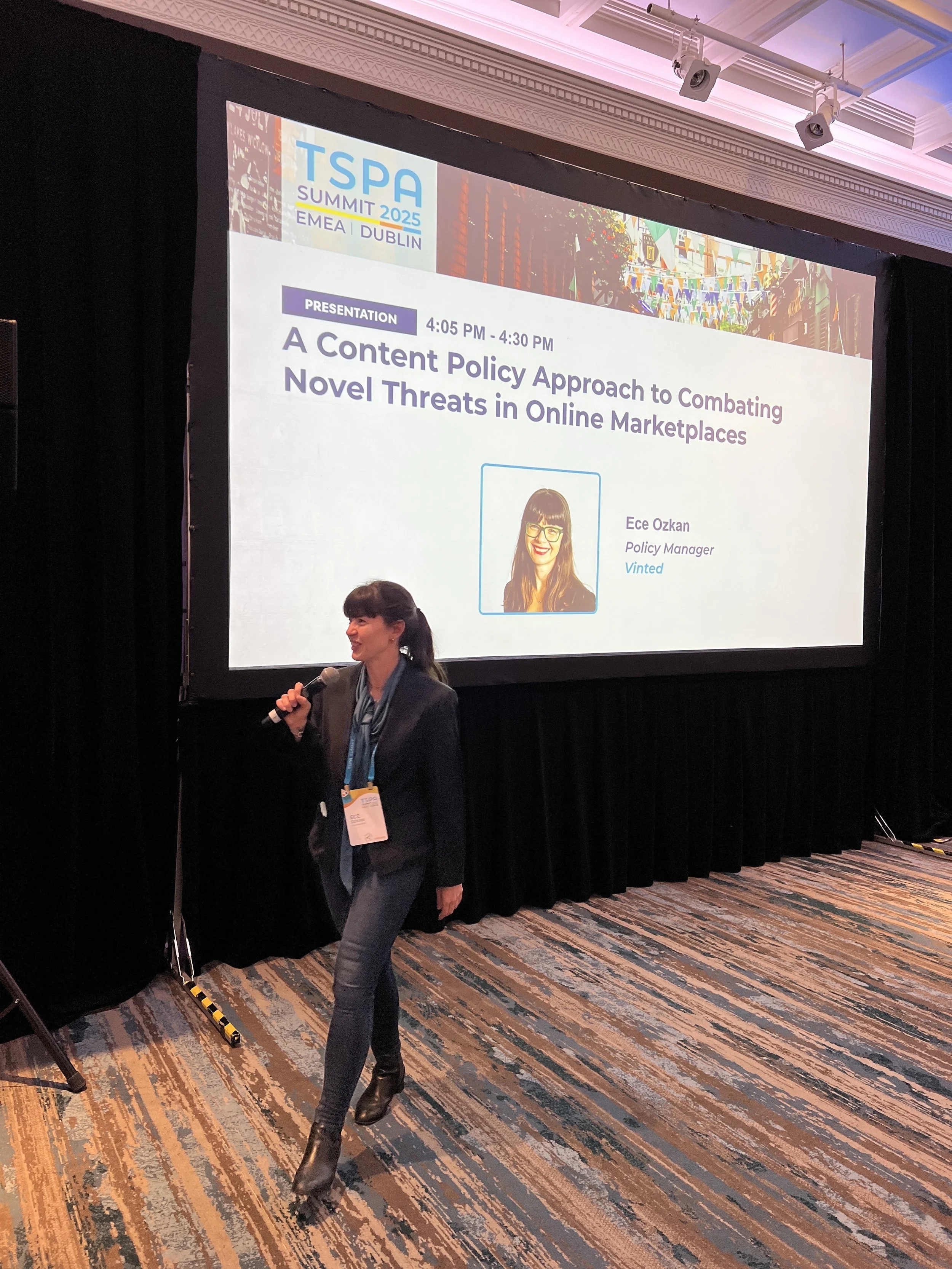 A woman walking past a large screen displaying a presentation slide at the TSPA Summit 2025 in Dublin. The slide features a photo of a smiling woman with glasses and the name Ece Ozkan, Policy Manager at Vinted, along with the presentation title: 'A Content Policy Approach to Combating Novel Threats in Online Marketplaces.' The conference room has a black curtain backdrop and a colorful, patterned carpet.