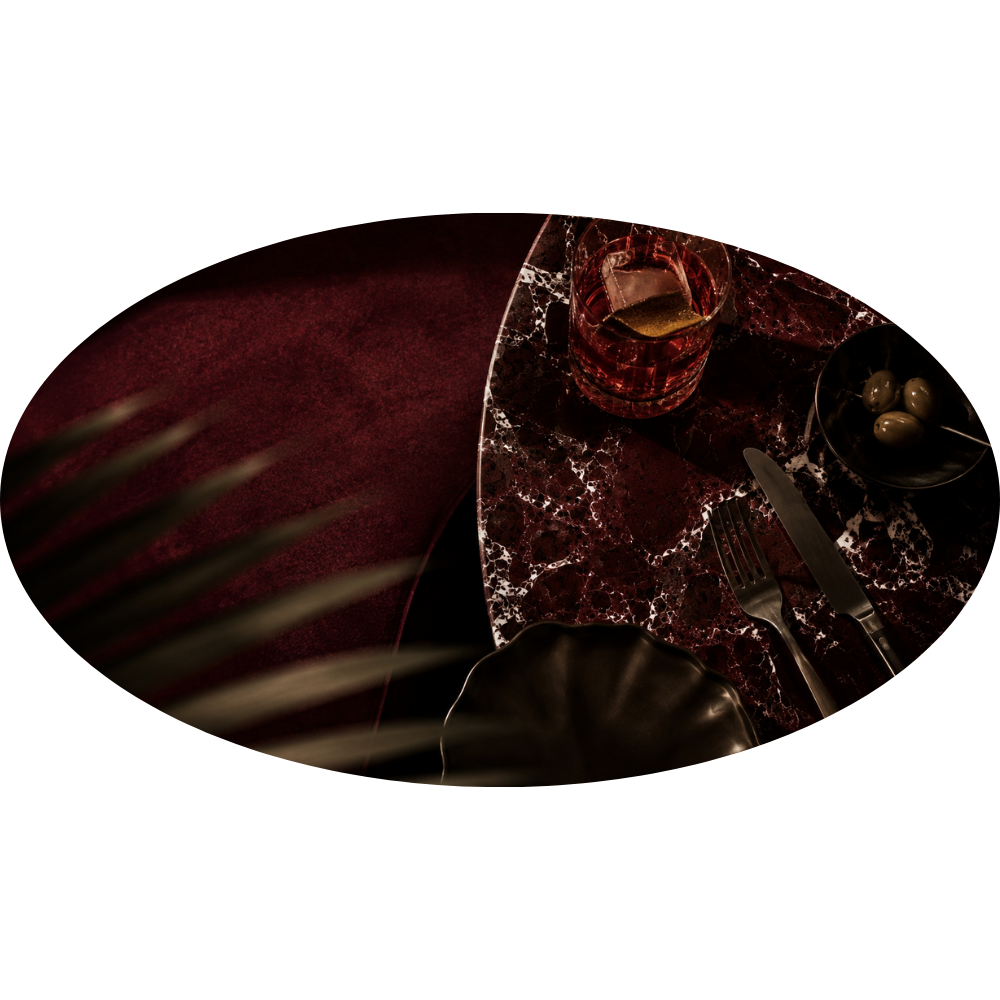 A table setting with a glass of drink, a bowl of green olives with toothpick, fork, knife, a dark plate, and a marble-patterned tablecloth, set on a red velvet table with dark background.