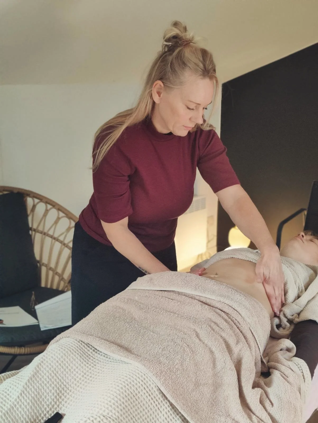 A woman in a burgundy shirt gives a massage to a person lying on a massage table, covered with a beige towel, in a cozy room.