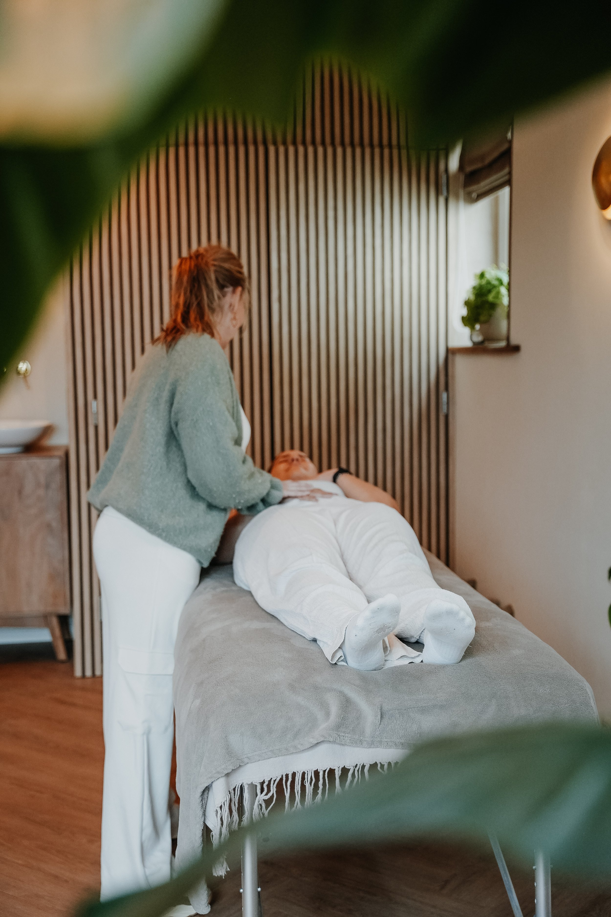 A massage therapist is performing a massage on a person lying face up on a massage table in a spa room with wooden floors and striped wooden wall paneling.
