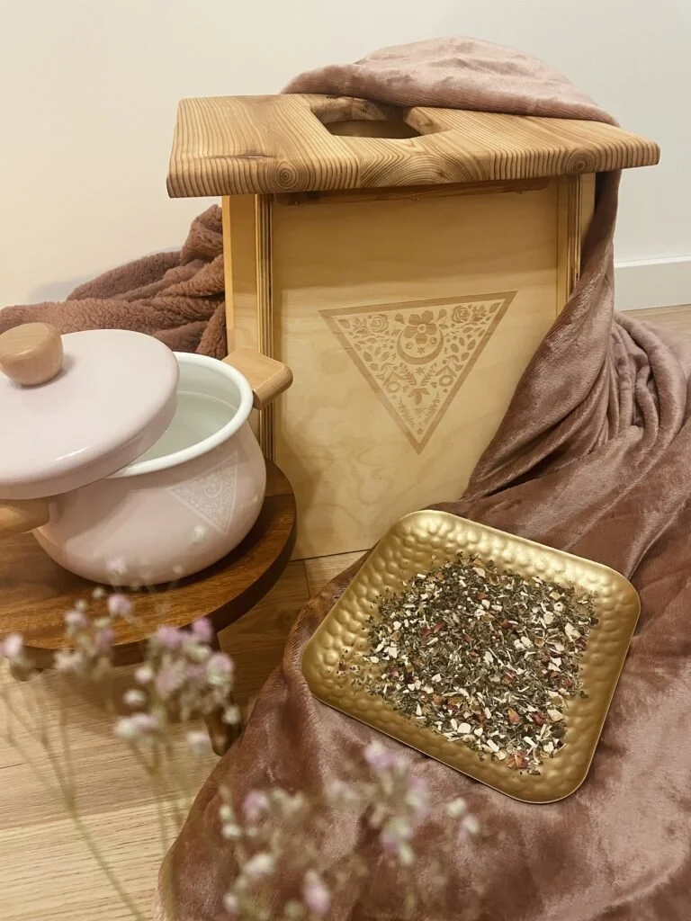 A wooden tea box with a floral design, a pink ceramic bowl with a lid, a golden textured tray filled with dried loose leaf tea, and soft pink and brown fabric draped around them, all placed on a wooden surface.