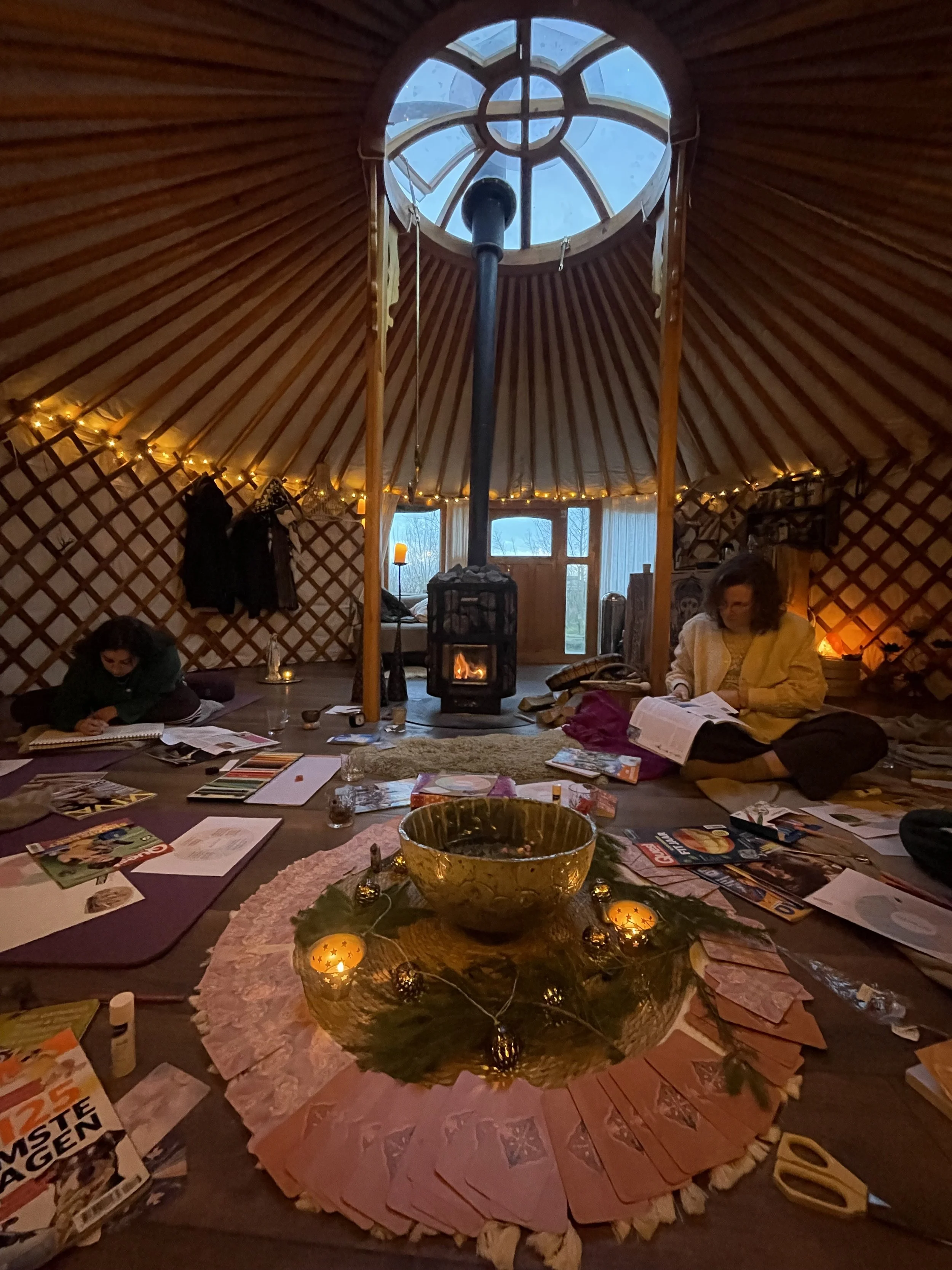 Inside a yurt with a central wood stove, people sitting on the floor, surrounded by books, magazines, and craft supplies, with a decorated table in the foreground and a skylight showing a blue sky.