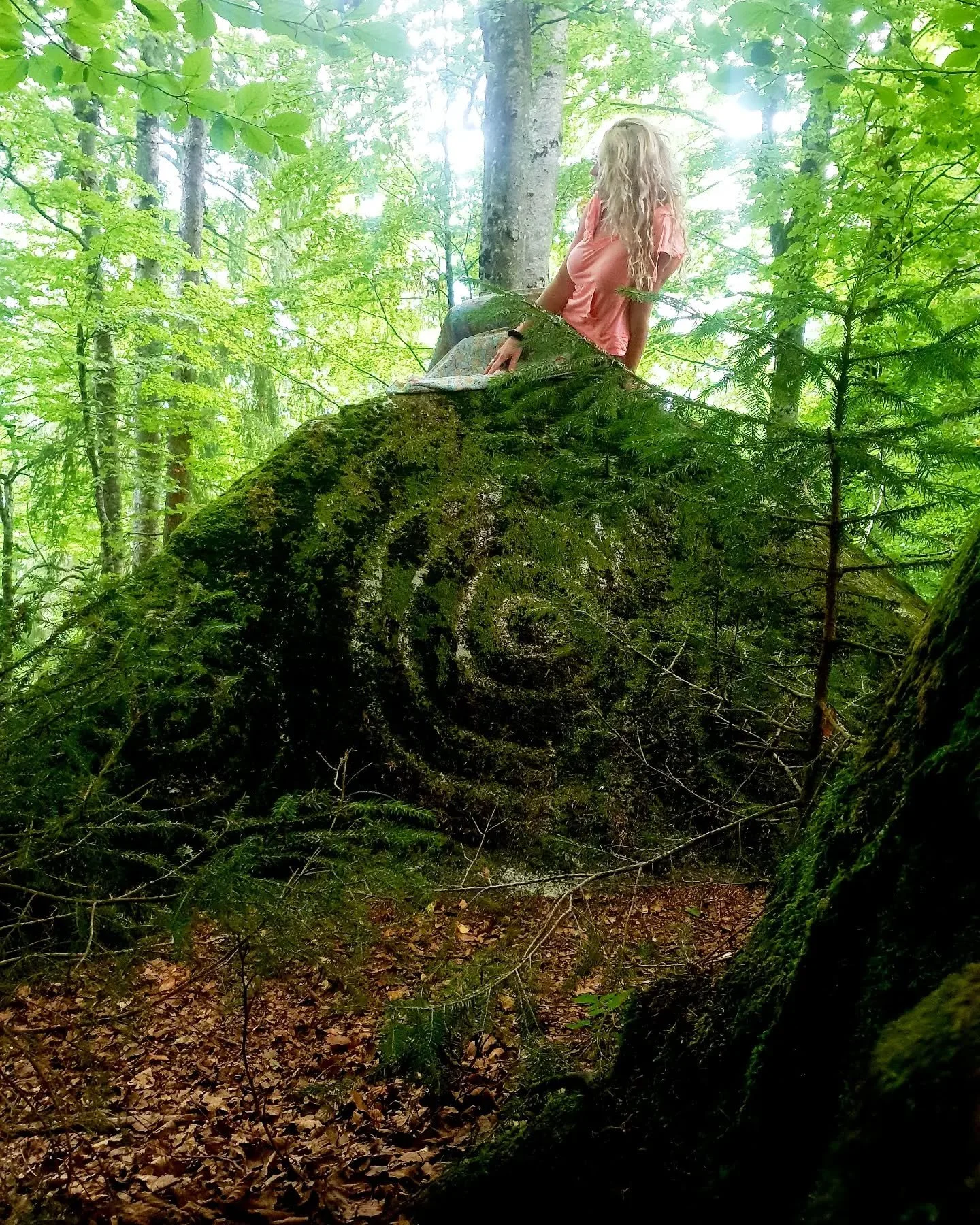 A woman with long blonde hair wearing a pink shirt sitting on a moss-covered fallen tree trunk in a dense green forest.
