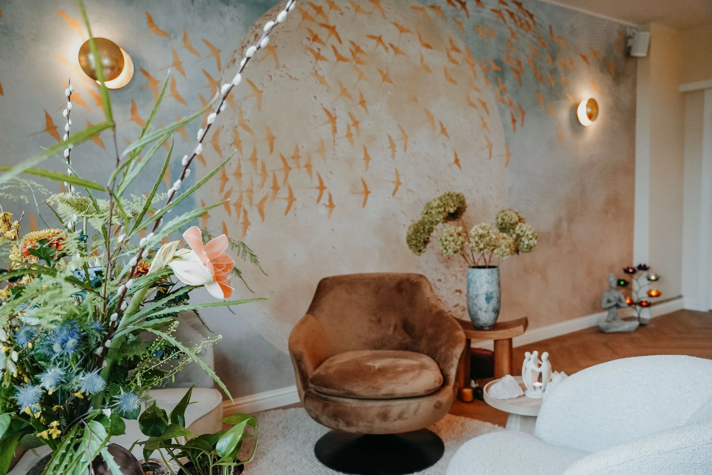 Living room with a beige velvet chair, a wooden side table, and an arrangement of flowers in a blue vase, with a textured wall decorated with orange bird silhouettes, two wall sconces, and a small Christmas tree with ornaments.