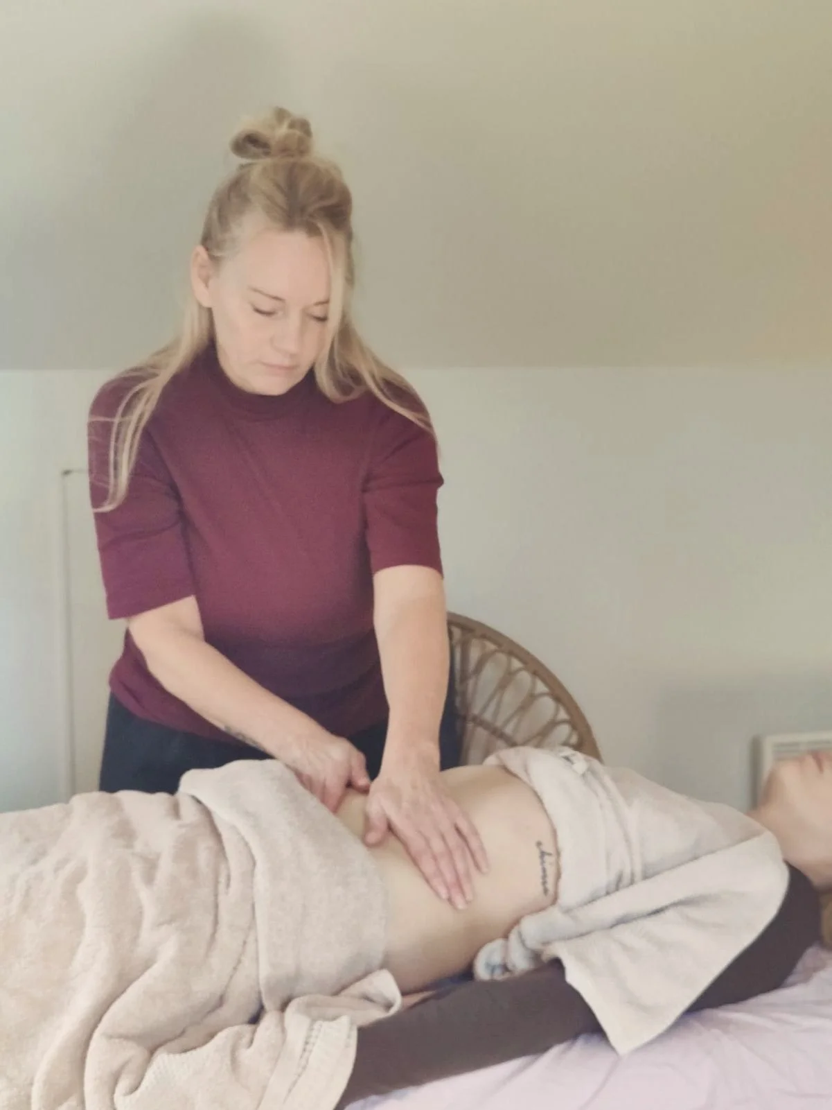 Woman giving a massage to a person lying face down on a massage table, wrapped in a towel, in a room with neutral-colored walls.