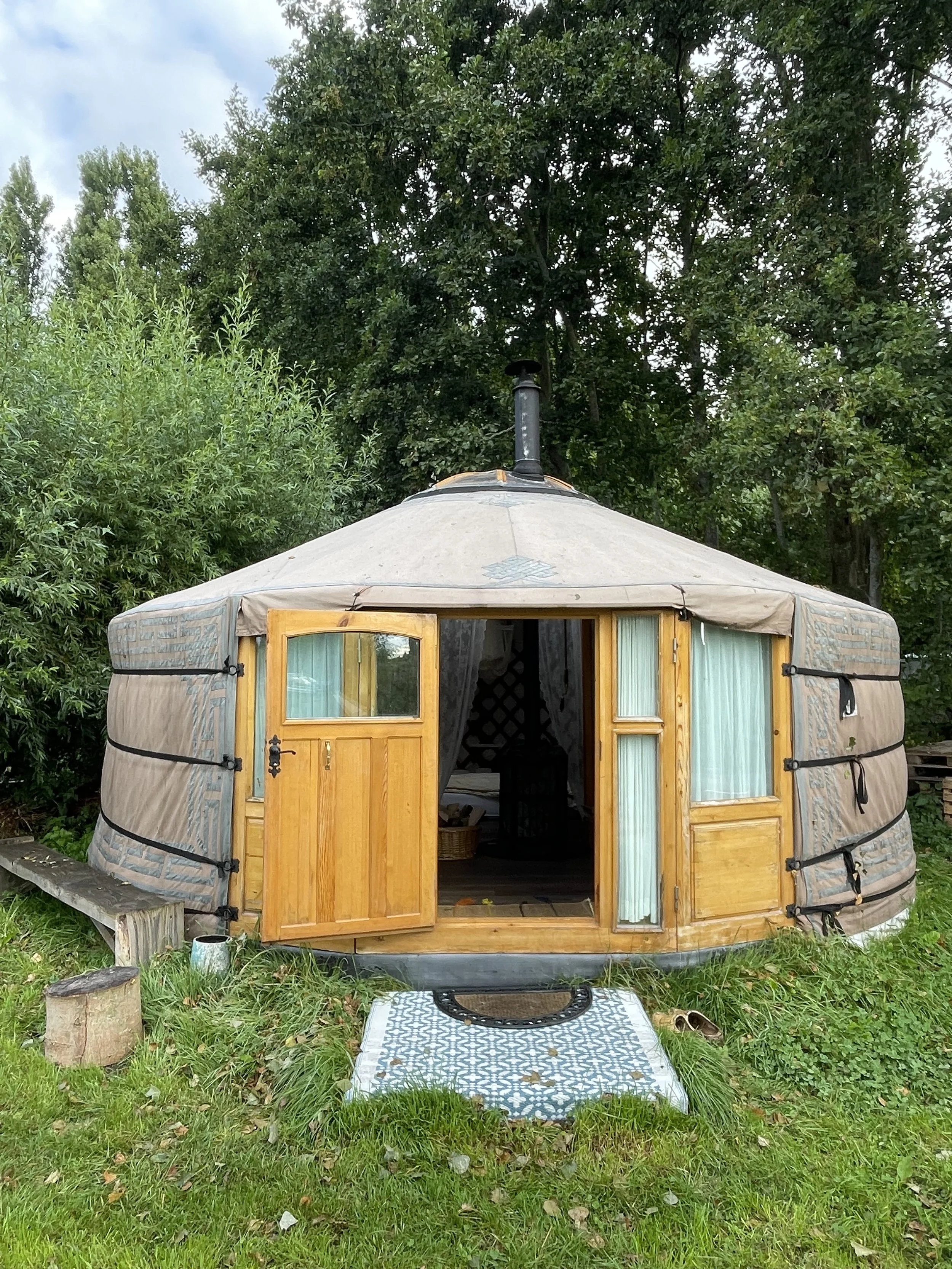 A round yurt with opened wooden door, sitting on a grassy area surrounded by trees, with a small welcome mat in front and a wood log on the left side.