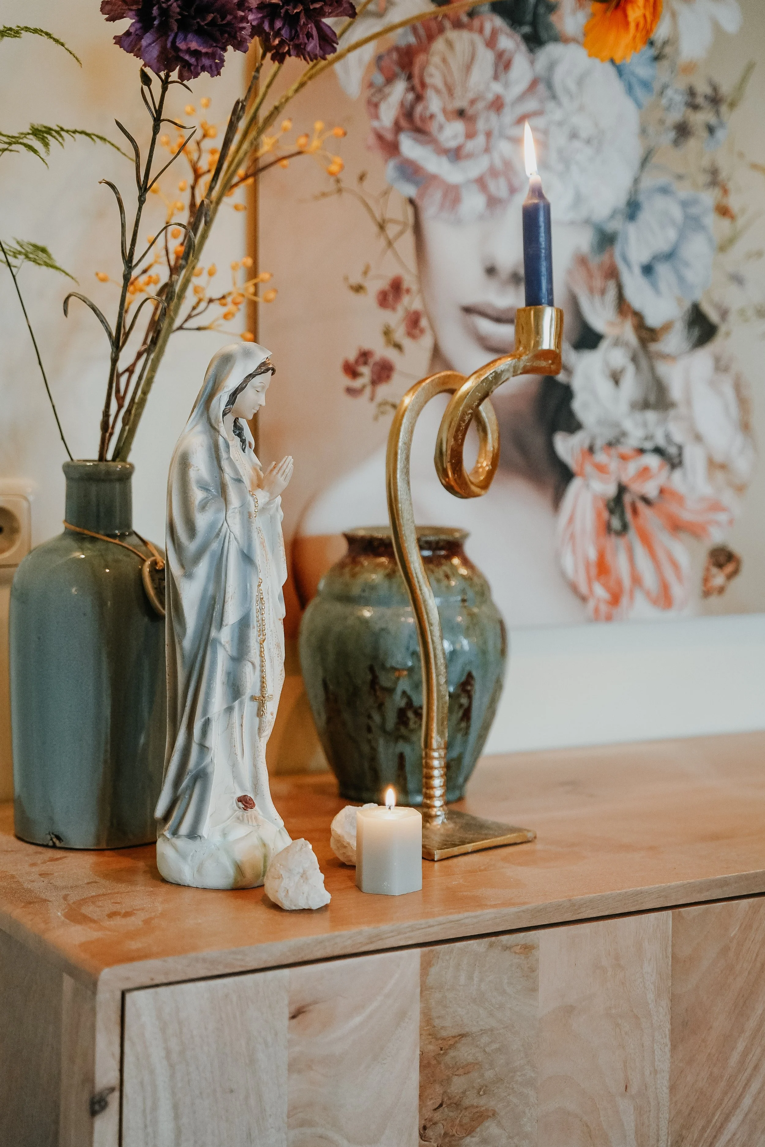 A decorative arrangement on a wooden surface featuring a figurine of the Virgin Mary, a lit candle in a green and gold candlestick, a small white candle, and various vases with dried and artificial flowers in the background, with a floral portrait of a woman on the wall.