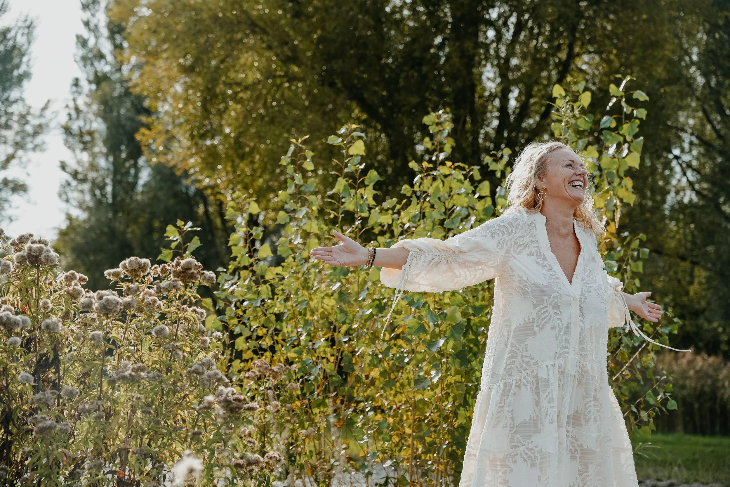 A woman in a white lace dress with outstretched arms smiling in a garden with green trees and flowers.