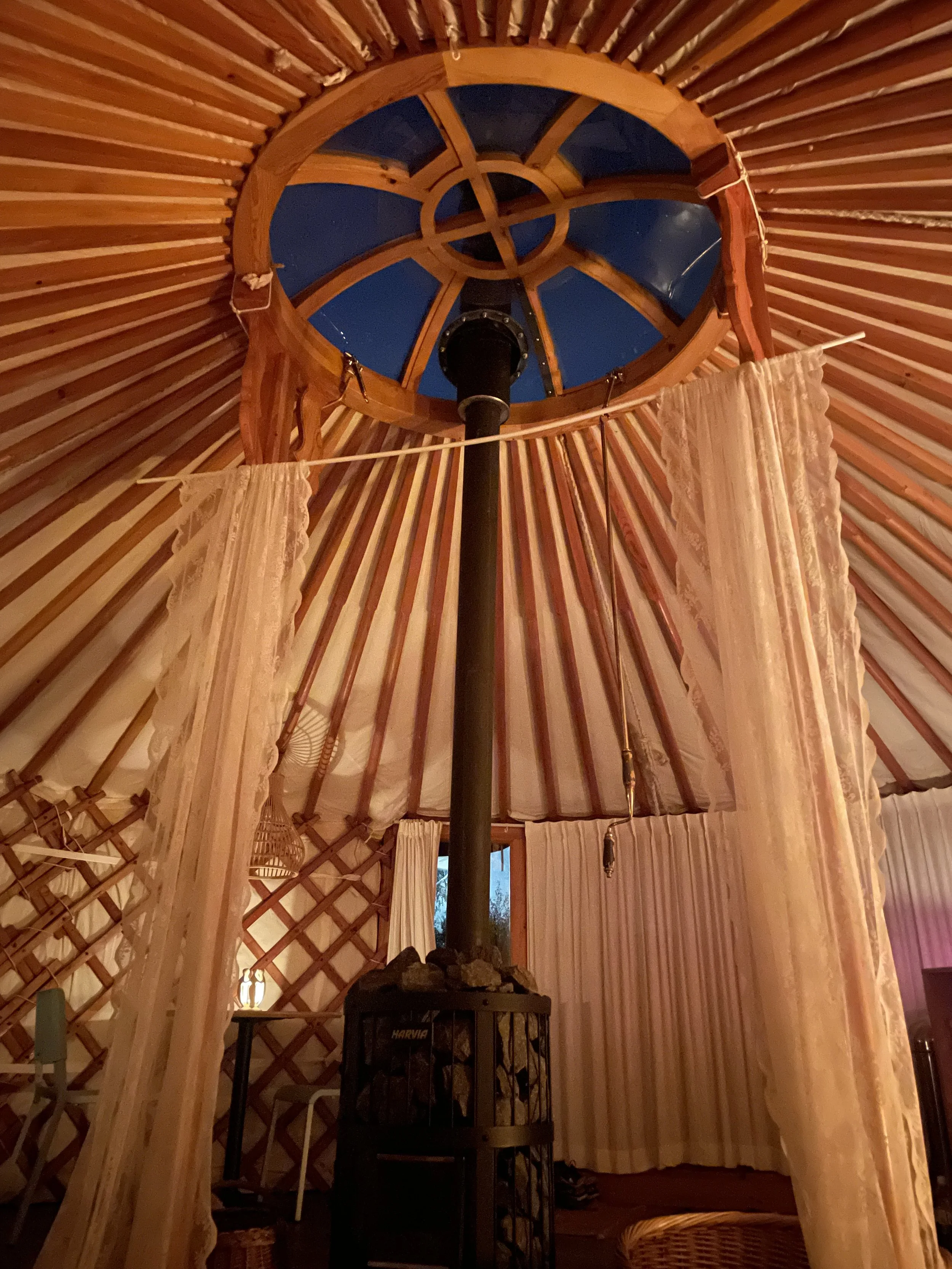 Interior view of a yurt with wooden lattice walls, fabric curtains, and a wooden ceiling. At the center, there is a black stove with rocks on top. A large circular skylight with wooden framing and blue sky outside is visible above.