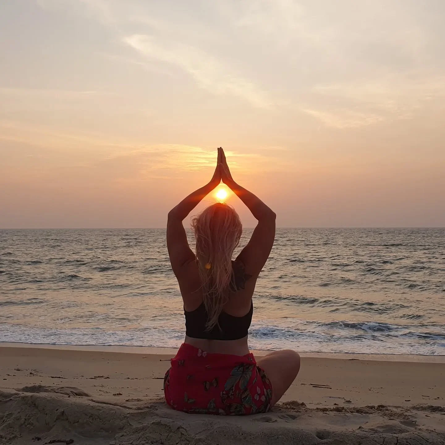 A woman practicing yoga on the beach during sunset with her hands in prayer position around the sun.
