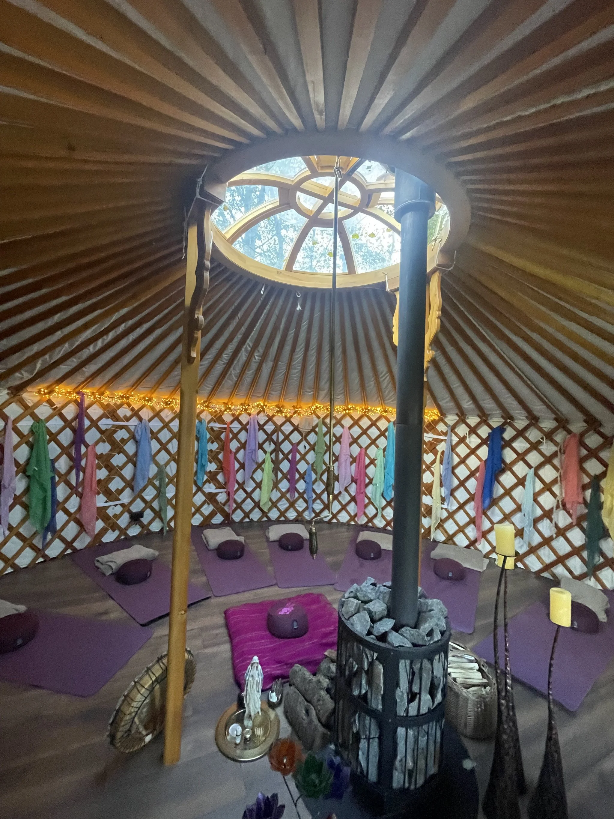 Inside a yurt with purple mats, pillows, and a purple blanket on the floor. A decorative fire pit in the center with rocks and candles surrounds it. Colorful fabric streamers hang on the lattice walls, and string lights provide warm lighting.