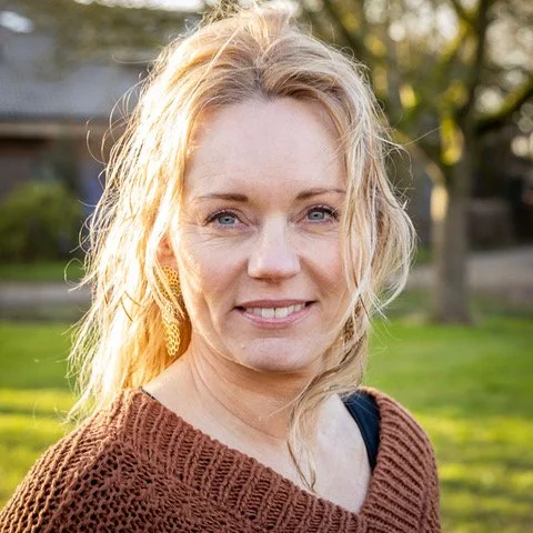 A woman with blonde hair and blue eyes smiling outdoors during golden hour.