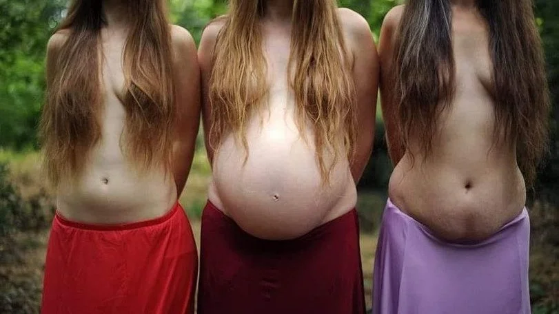 Three pregnant women standing outdoors with long hair, wearing skirts in red, maroon, and purple, with their midsections exposed.