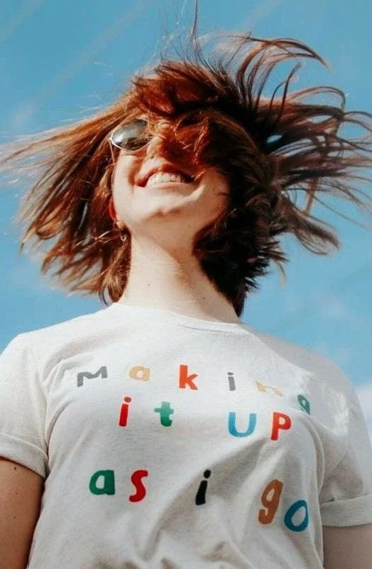 Una ragazza sorridente con i capelli mossi al vento, indossa una maglietta bianca con scritte colorate. Sullo sfondo un cielo azzurro.