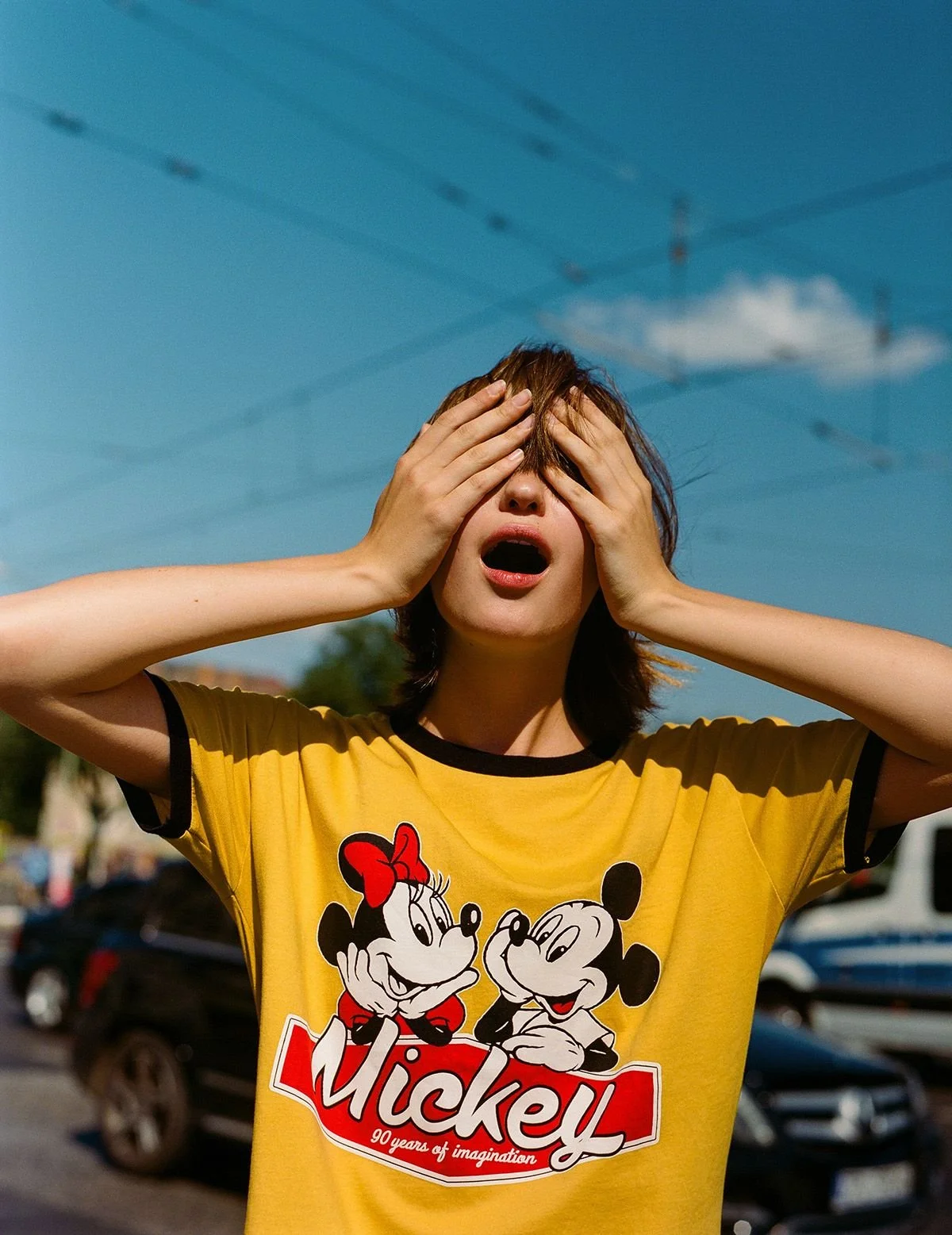 Una donna con una maglietta gialla con personaggi Disney, che si tiene la testa con le mani e ha un'espressione di shock o sorpresa, scatti in un giorno di sole con veicoli e alberi sullo sfondo.