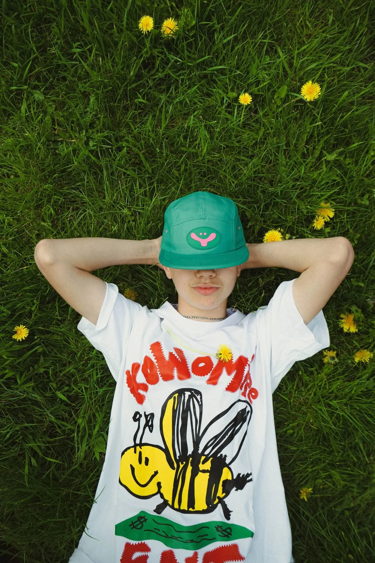 Ragazzo sdraiato sull'erba con fiori gialli, indossa una maglietta bianca con disegni e una cappellino verde con sorriso