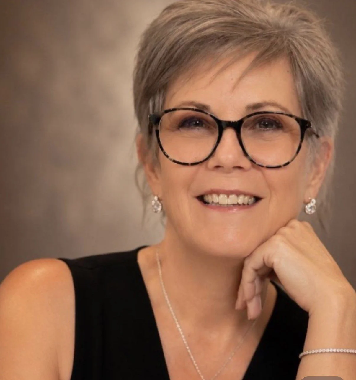 Paula, a smiling woman with short hair, wearing glasses, earrings, and a black top, resting her chin on her hand.