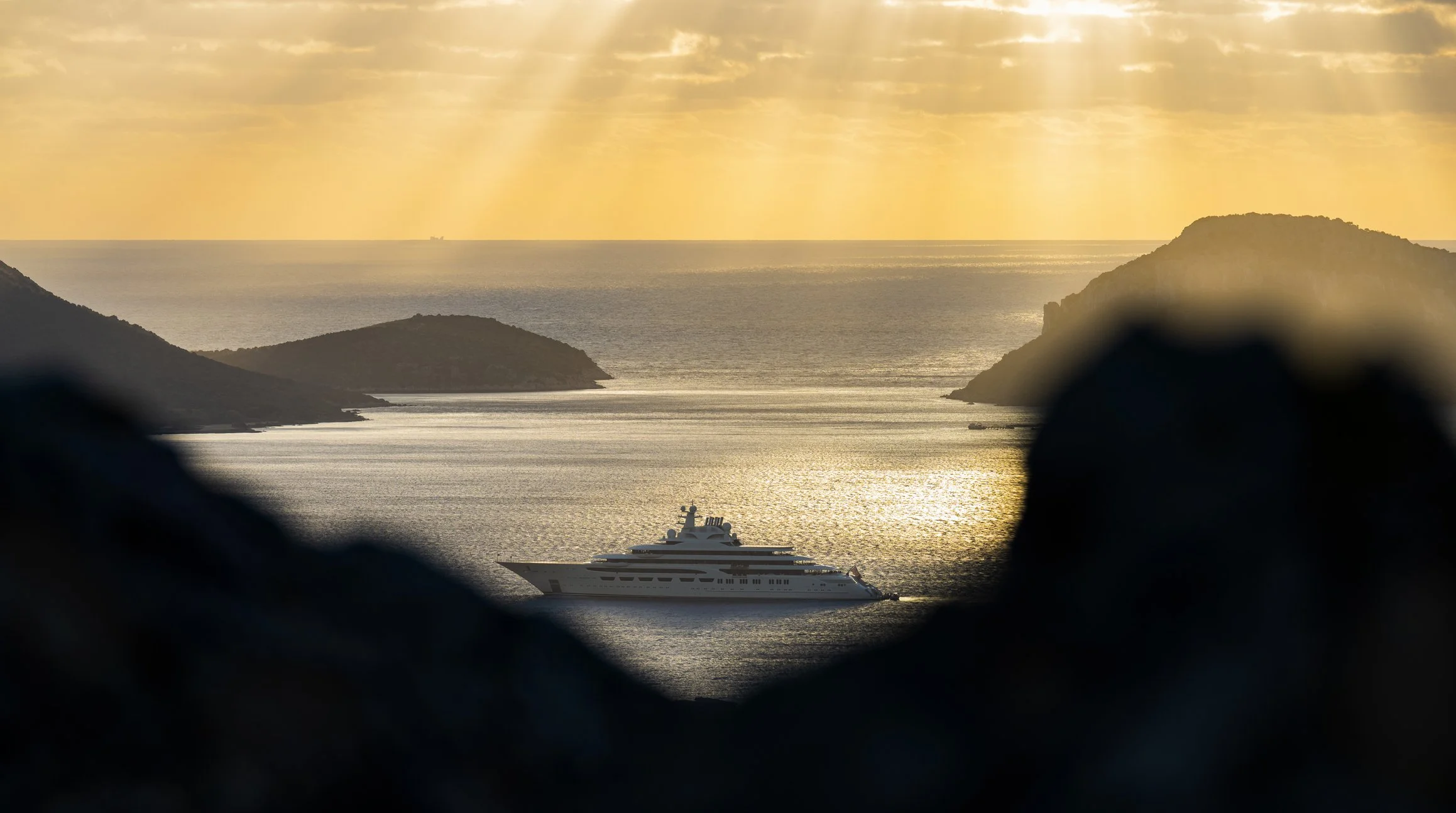 A luxury yacht sailing through the ocean at sunset, with islands and a ship visible in the distance, framed by dark rocks in the foreground.