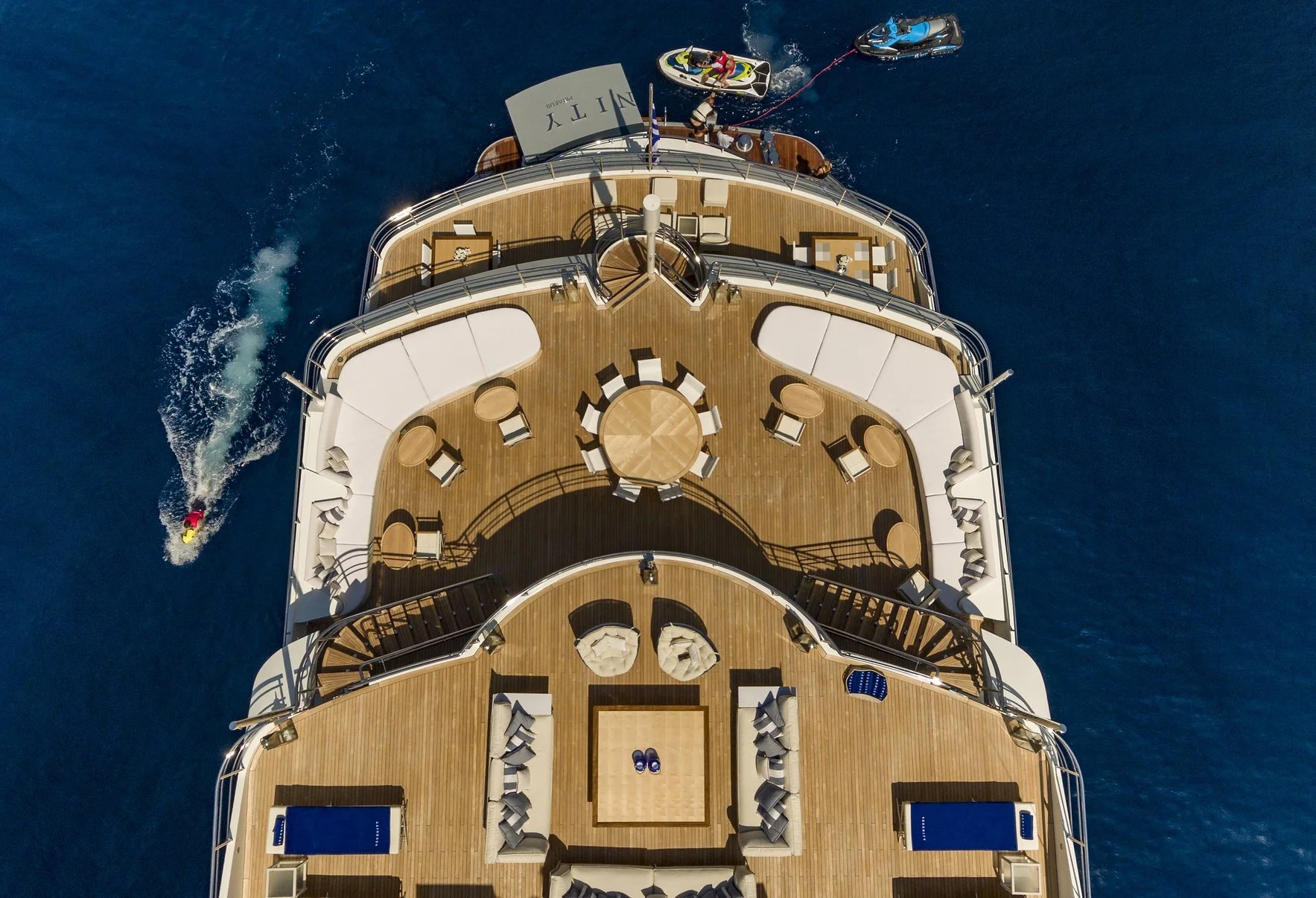 Top-down aerial view of a large yacht with a spacious deck featuring seating areas, tables, lounge chairs, and a person waterskiing behind it in the ocean.
