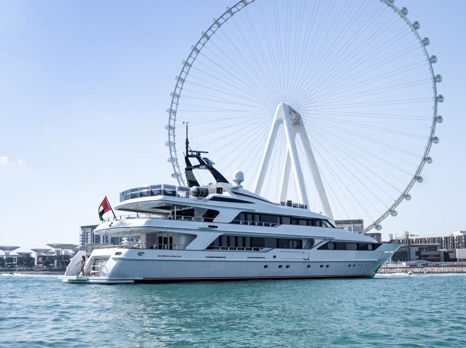 A large white yacht is docked near a city waterfront with a giant Ferris wheel in the background. The yacht has multiple decks and a flag at the stern.