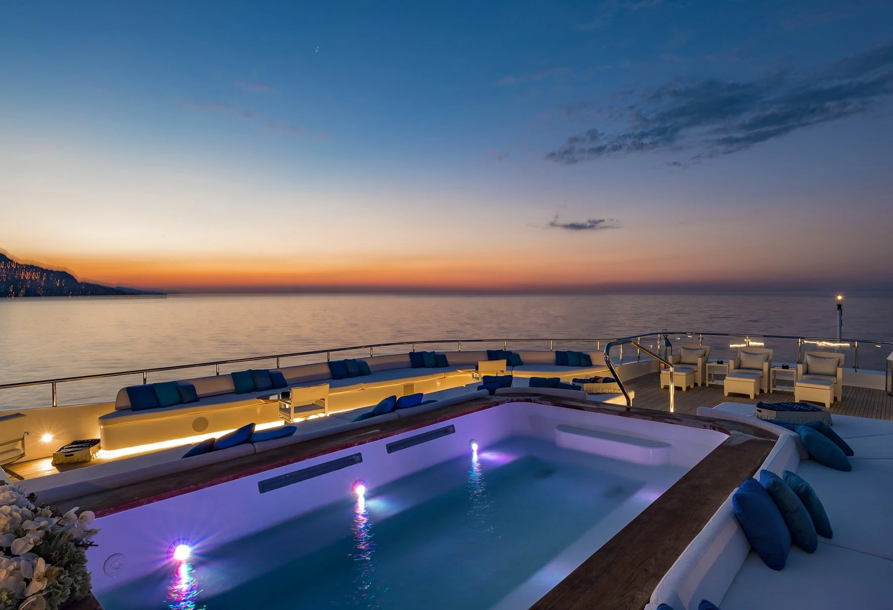 Luxury yacht deck at sunset with a small illuminated hot tub, seating areas with cushions, and a view of the calm ocean and colorful sky.