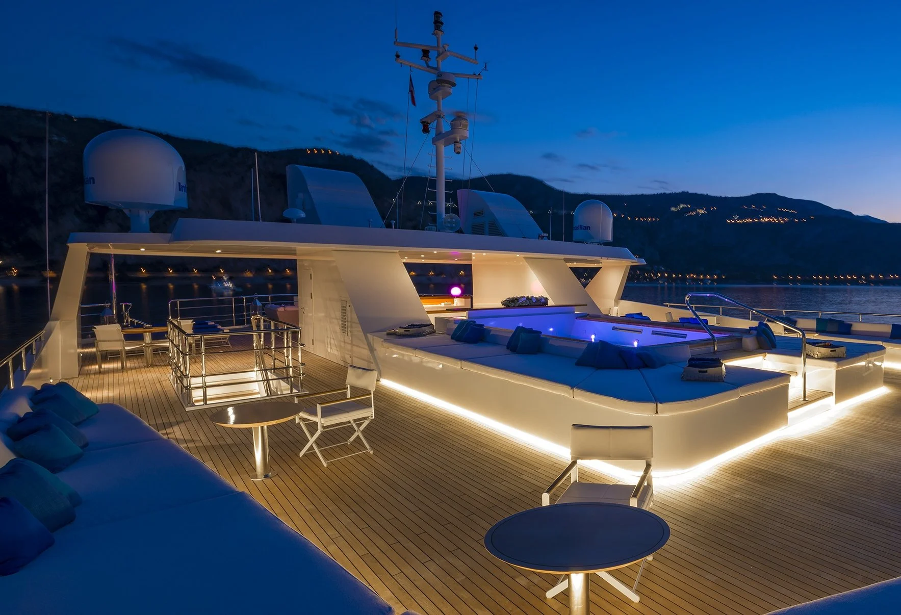 Luxury yacht deck at dusk with outdoor seating, lounge area, and illuminated lighting, overlooking a water body with hills in the background.