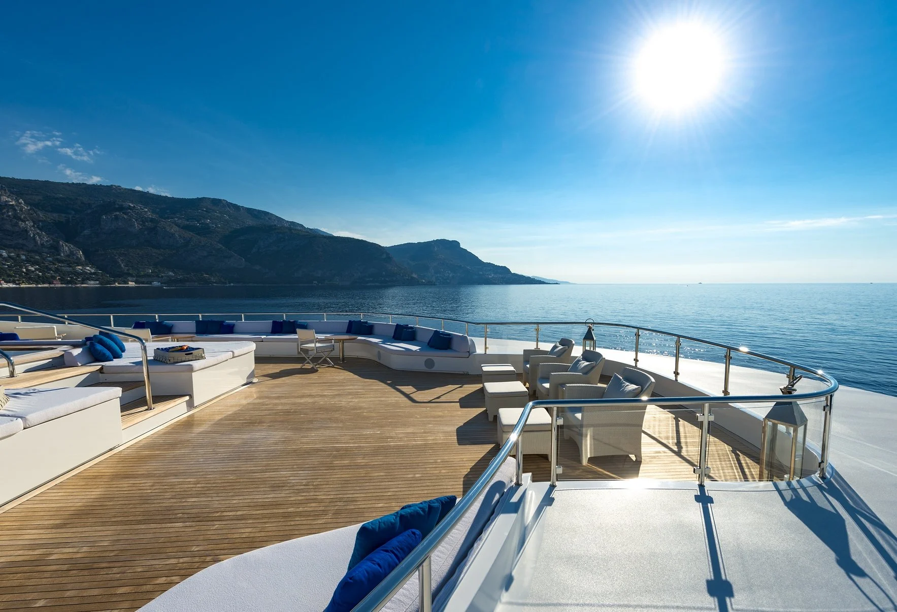 Luxury yacht deck with white seating and blue pillows, overlooking a calm ocean with mountains in the background and a bright sun in the clear blue sky.