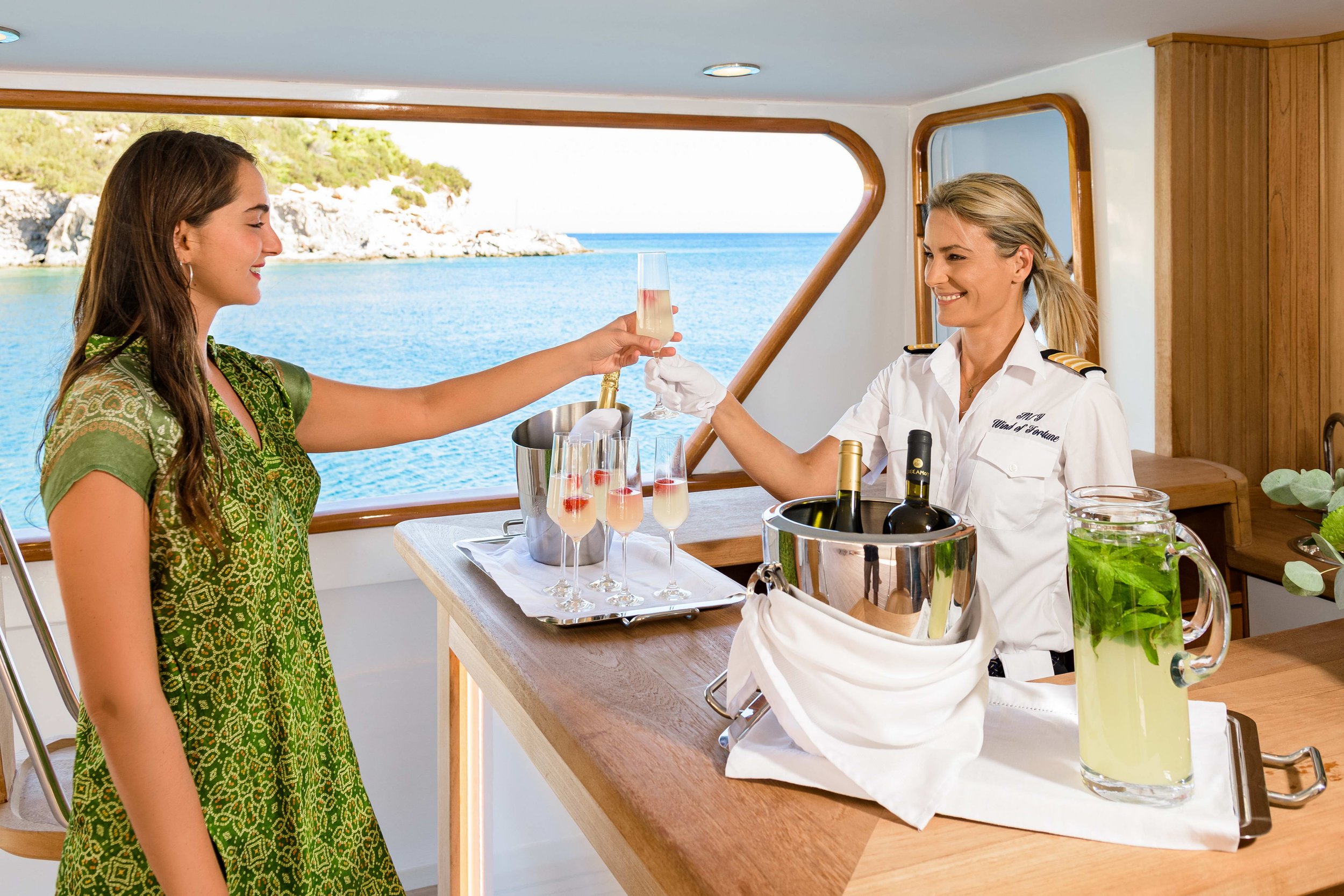 A woman in a green dress receives a glass of champagne from a woman in a white uniform on a boat with a view of the ocean and coastline outside the window.