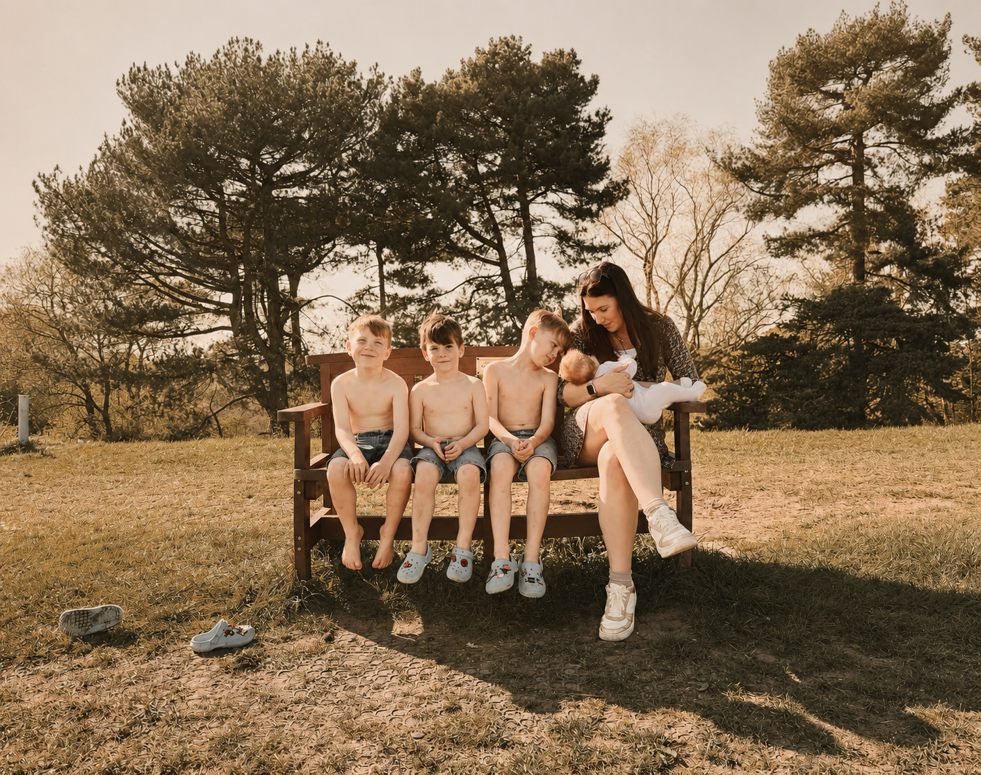 A woman sitting on a park bench surrounded by four young boys in the sun, with trees in the background.