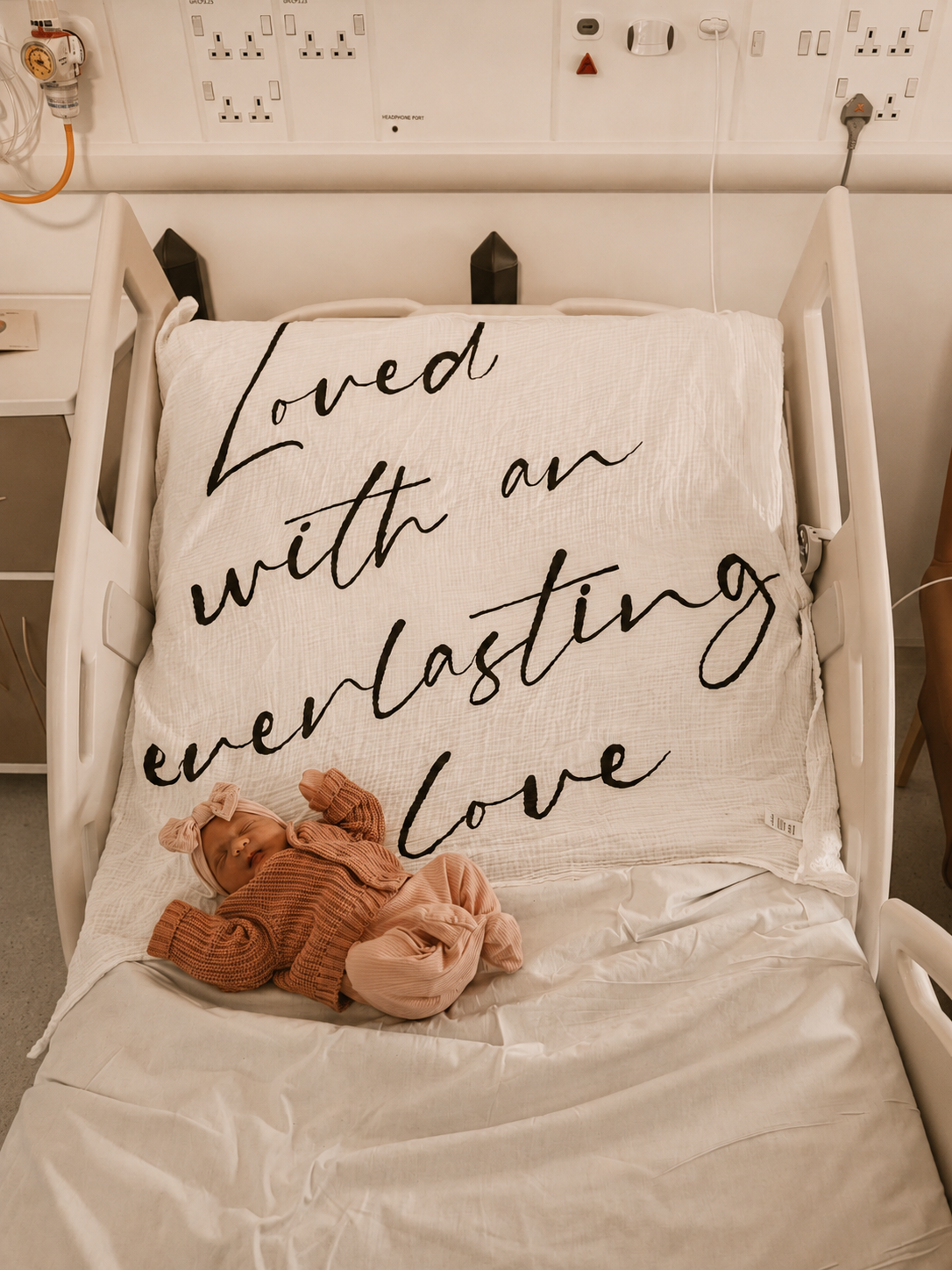 A hospital bed with a large pillow that has black cursive text reading "Loved with an everlasting love." A sleeping baby wearing pink and brown clothing, including a headband with a bow, lies on the bed.