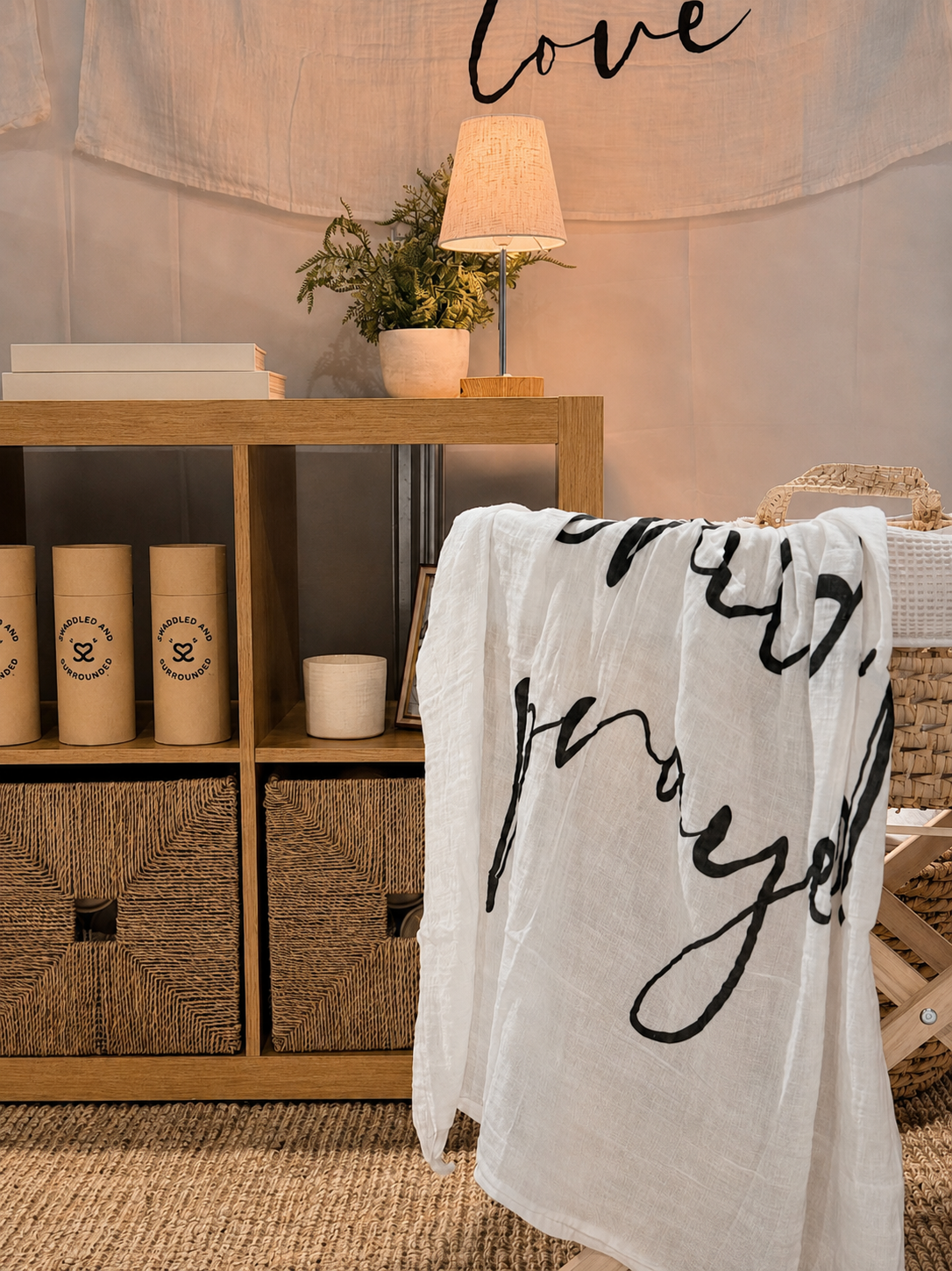 A cozy interior scene featuring a wooden console table, a table lamp, a potted plant, and a white cloth with black cursive writing hanging over a basket and ladder. The wall above has the word 'love' written in black.