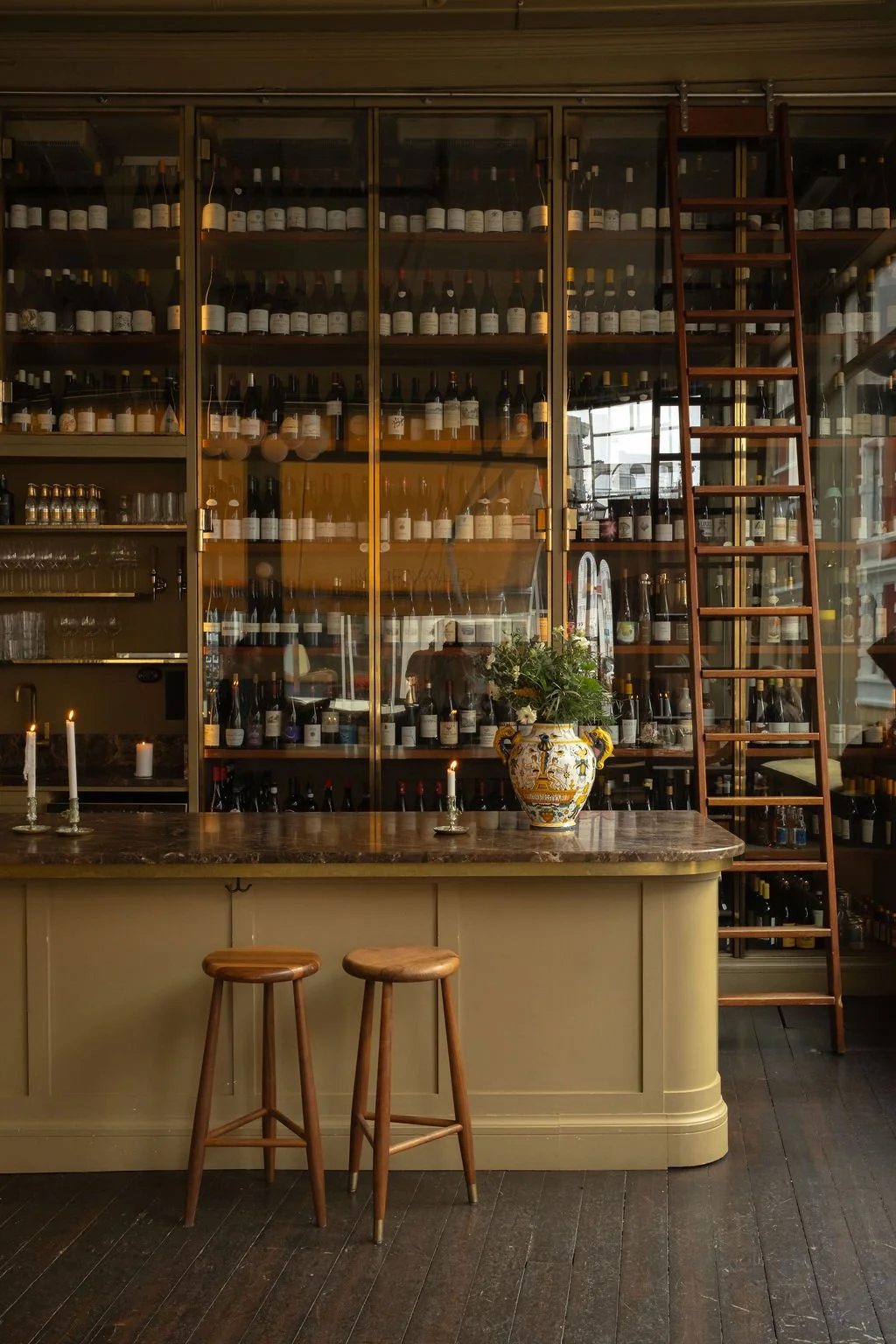 Interiør av en vinbar med en bar, en glasskap fylt med vinflasker, en ladder, en vase med blomster, og to barkrakker.