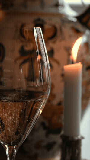 Et glass med champagne, en hvit lysende stearinlys og en person med blomstermønster i bakgrunnen.