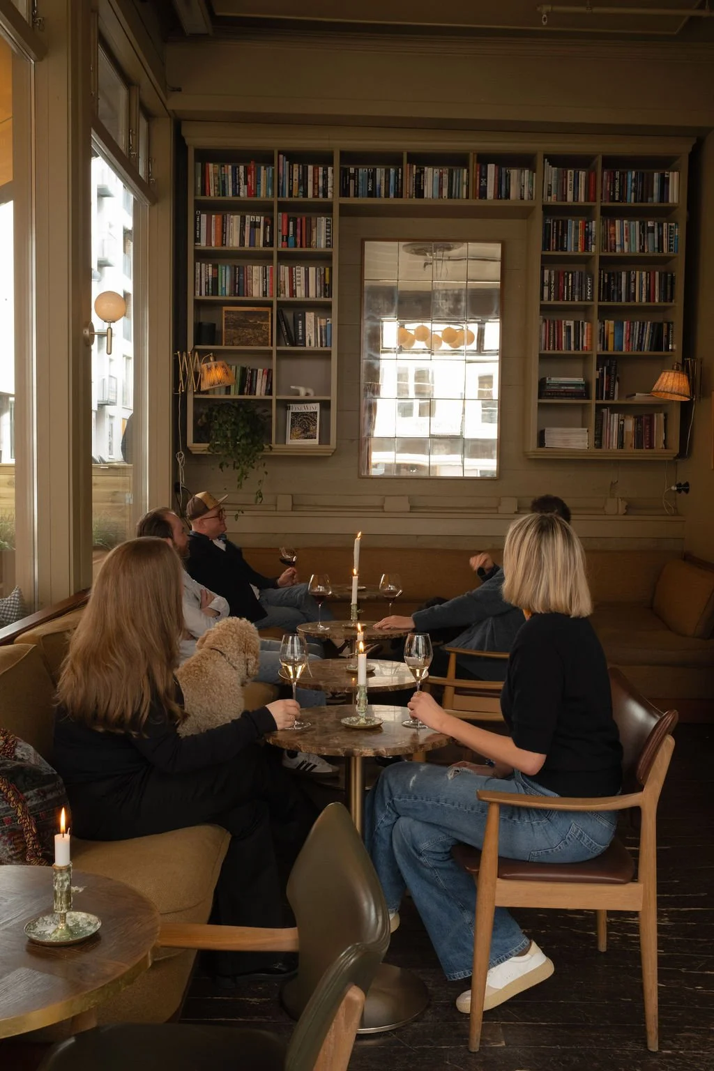 Innendørs scene med fem personer som sitter rundt et bord med stearinlys, vin og en hund. Rikelig med bokhyller og store vinduer i bakgrunnen.