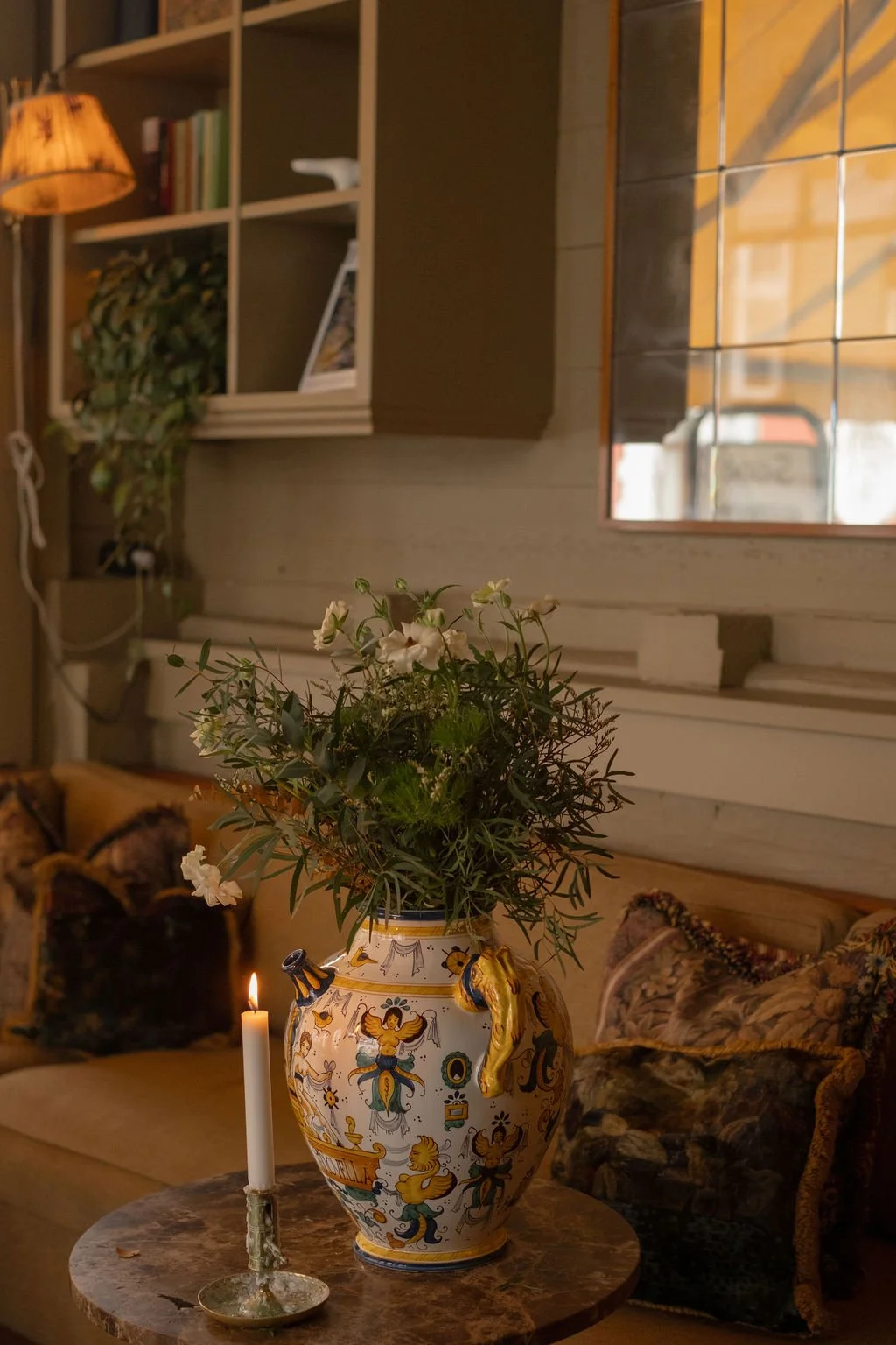 En dekorativ vase med grønne planter og hvite blomster, plassert på et rundt trebord ved siden av et hvitt lys i en liten brikker, i et koselig rom med en sofa og dekorative puter, samt hyller med bøker og et plantestell i bakgrunnen.