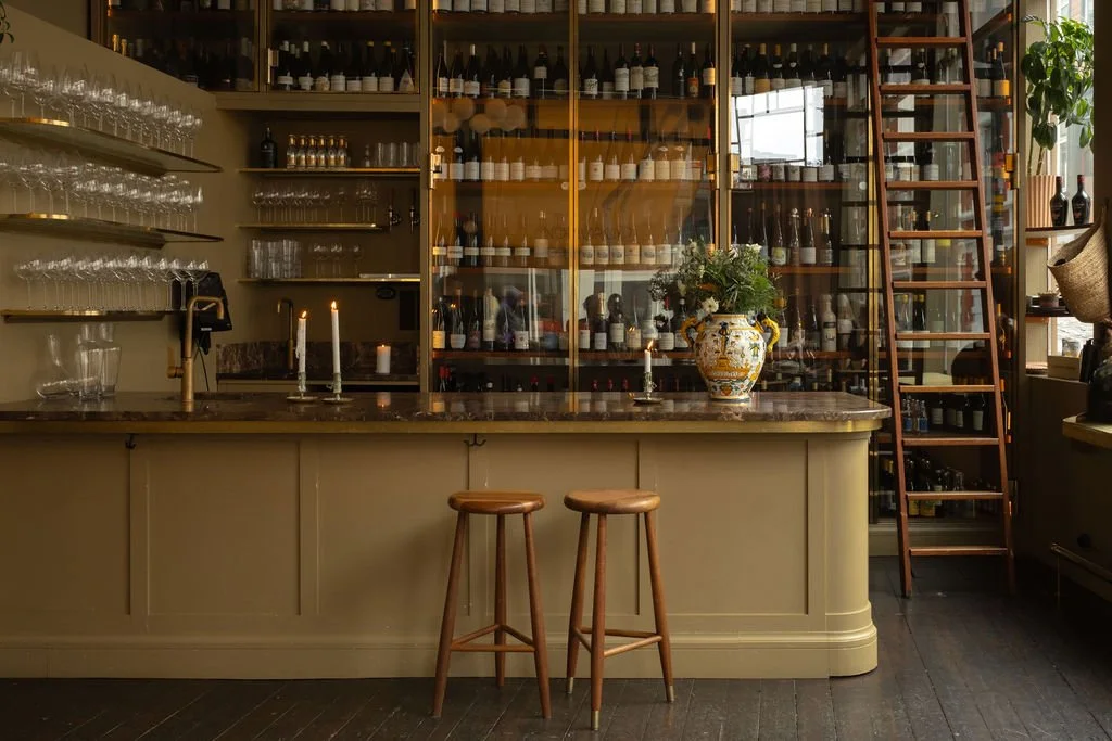 Interiør av en bar eller kafé med hyller fylt med vin- og drikkeflasker, et bardisk med to stoler, lysestaker, og en stor vase med blomster.