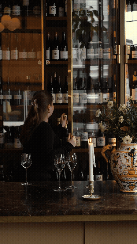 En person står bak baren og åpner en vinflaske, omgitt av tomme vin- og cocktailglass og dekorasjoner i et sofistikkert restauranten eller baren.