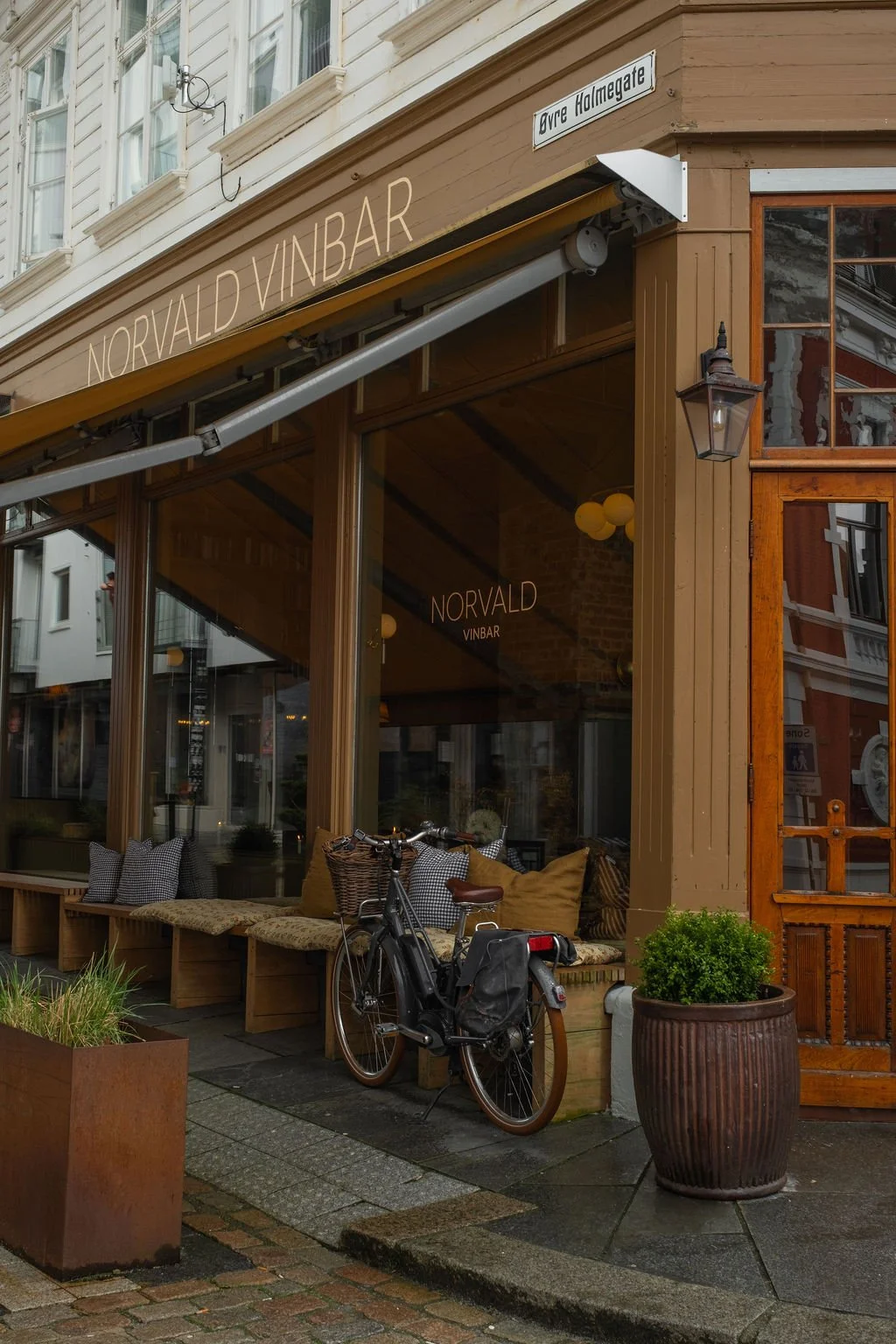 Utenfor en vinbar med navn Norvald Vinbar på Øvre Holmegate i Oslo. Det er en sykkel parkert utenfor, og det er store vinduer med sitteplasser innendørs. Det er også en stor blomsterpotte med grøntanlegg og en lampe utendørs.