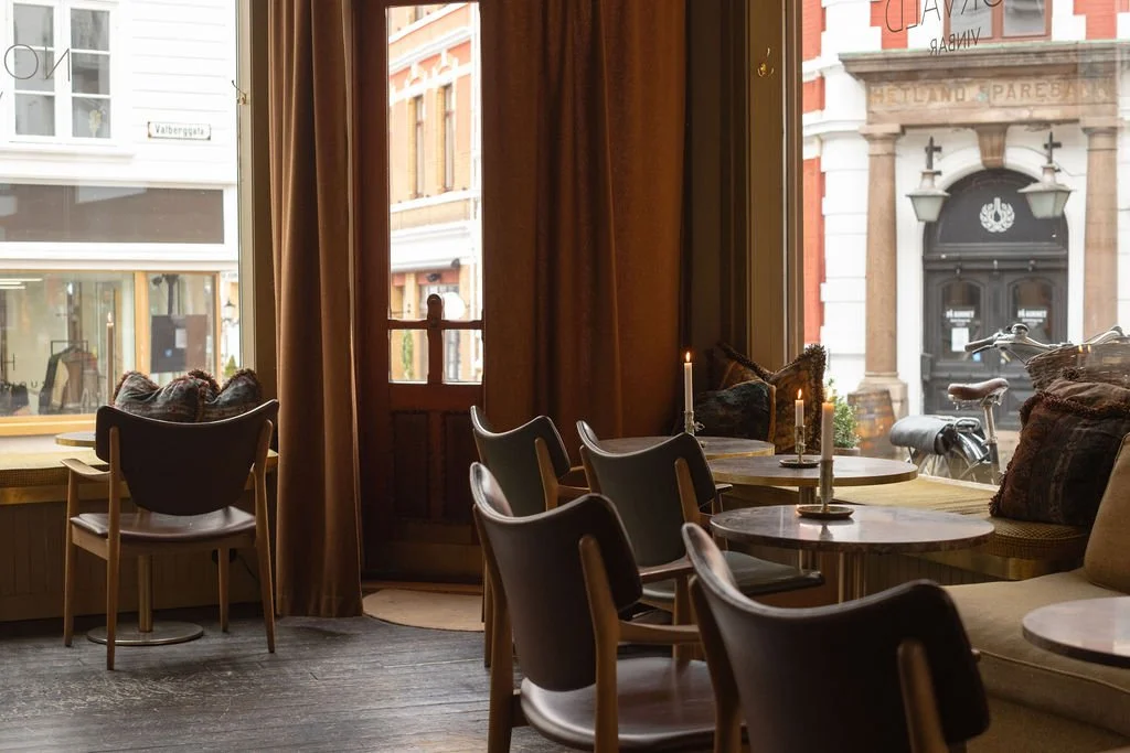 Innendørs kafé med tre bord, noen med stearinlys og puter, vinduer som viser en gate med gamle bygninger utenfor.