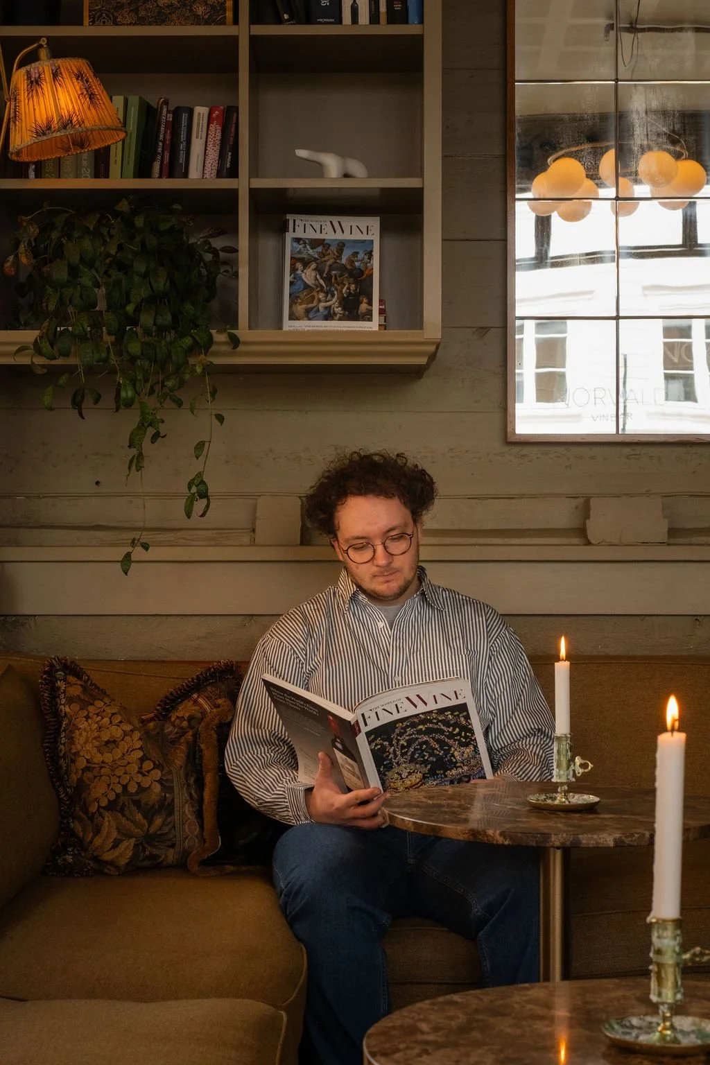 En mann med briller som leser en magasin kalt 'Fine Wine' mens han sitter på en kafé, med tente lys på bordet foran ham. Bak ham er det en bokhylle med bøker, planter og dekorative gjenstander.