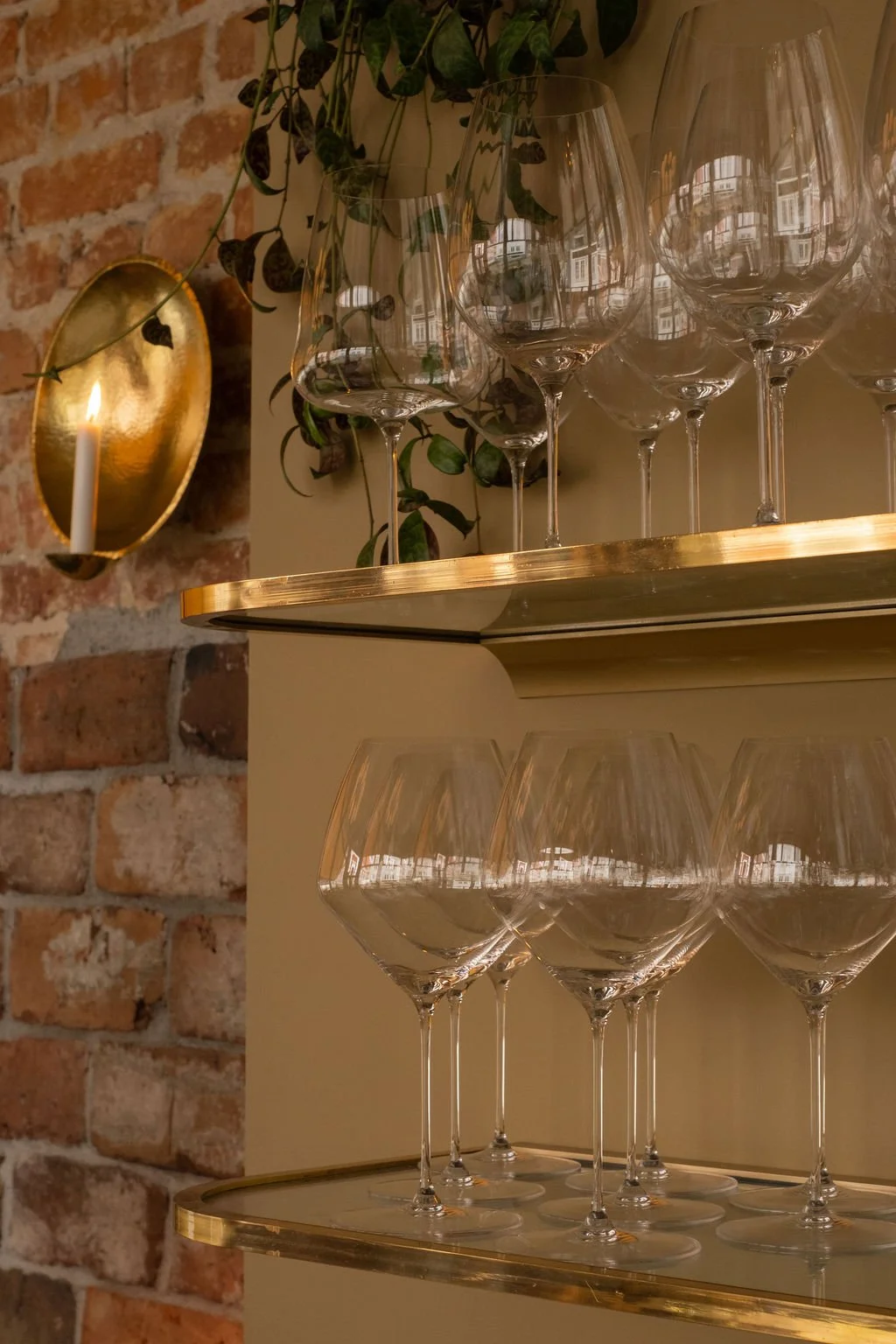 Tolv glass vinglass på en gullfarget hylle med utsikt over vinduer, og en vegg av murstein med en vegglampe med lys.