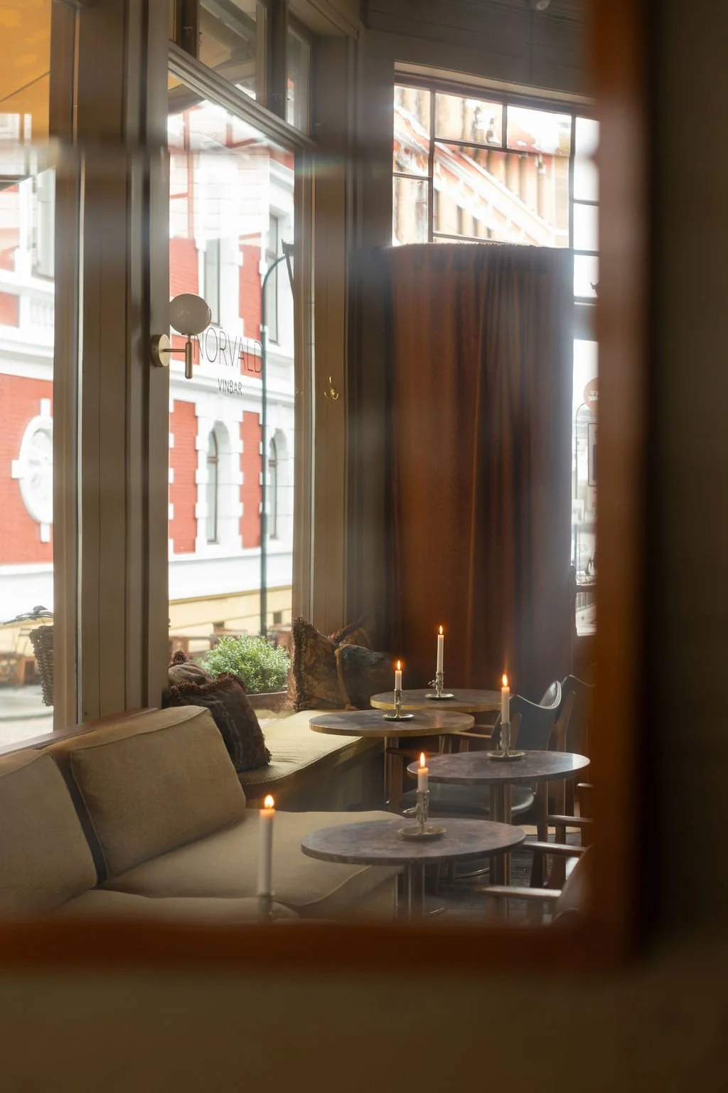 Interiør av en kafé med puter, små bord med tente stearinlys, og store vinduer som viser en bygning utenfor.