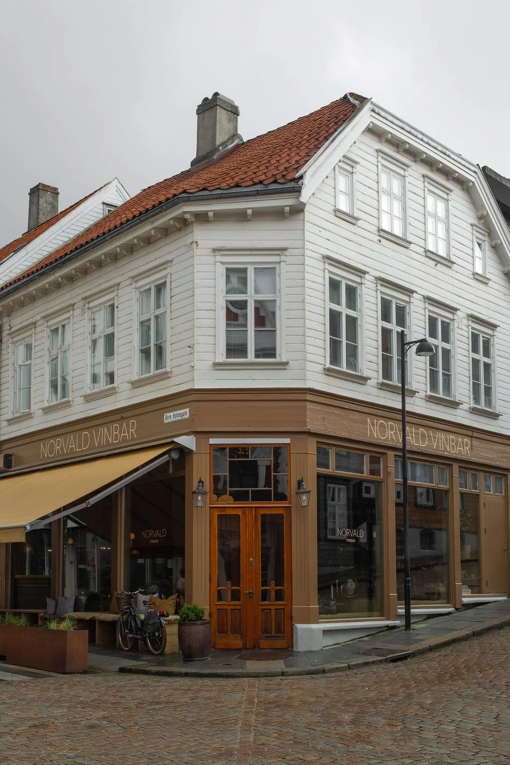 Tverrbygning med hvit trefasade, rød takstein, og skilt som sier Norvald Vinbar. Nedre del av bygningen er innredet som kafé eller vinbar med treinngang og vinduer. Utendørs sitteplass og gatebelysning. Skummelt overskyet vær.