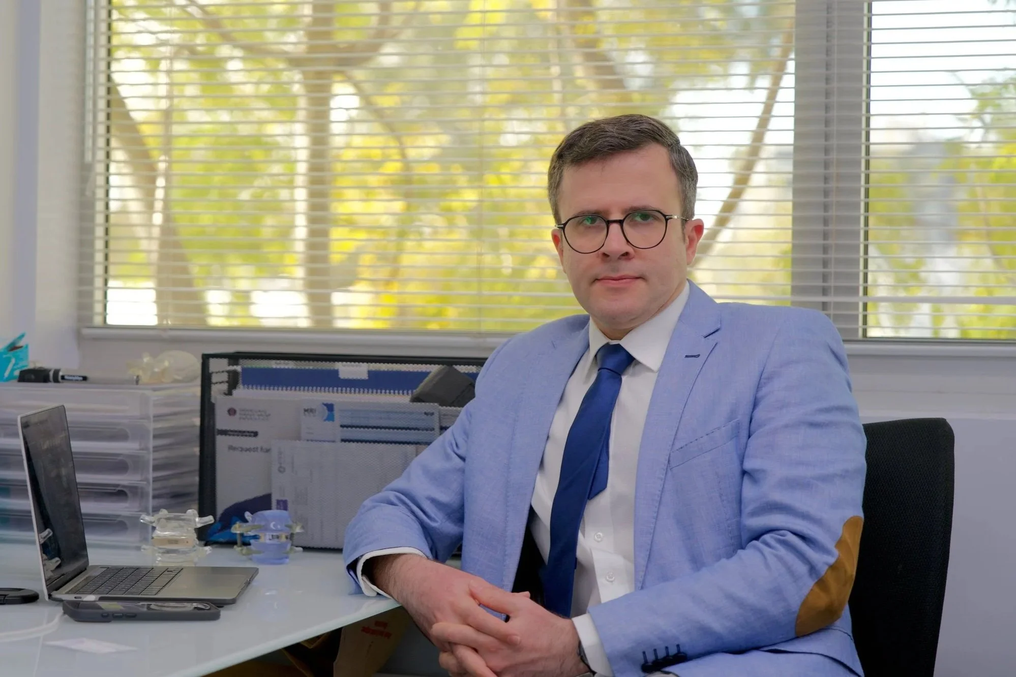 A man with brown hair, glasses, wearing a light blue suit and dark blue tie, sitting at an office desk in front of a window with blinds, in a professional office setting.